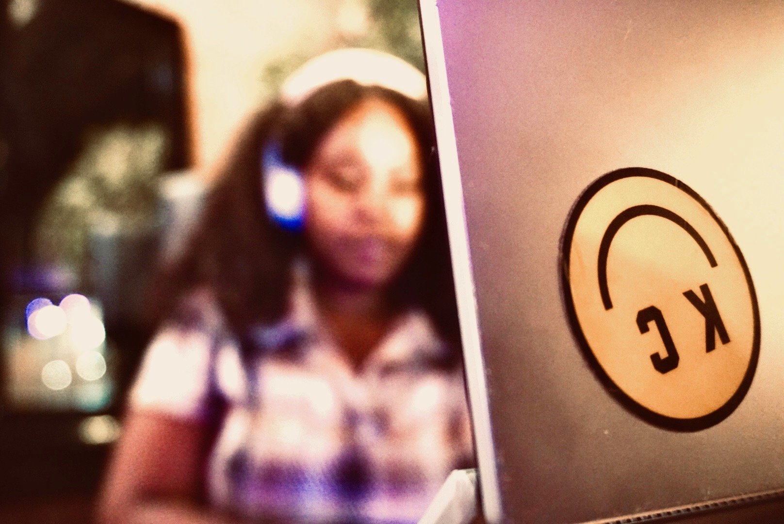 A woman wearing headphones is out of focus in the background, while a close-up of a laptop with a sticker that reads 'KLC' is in the foreground.