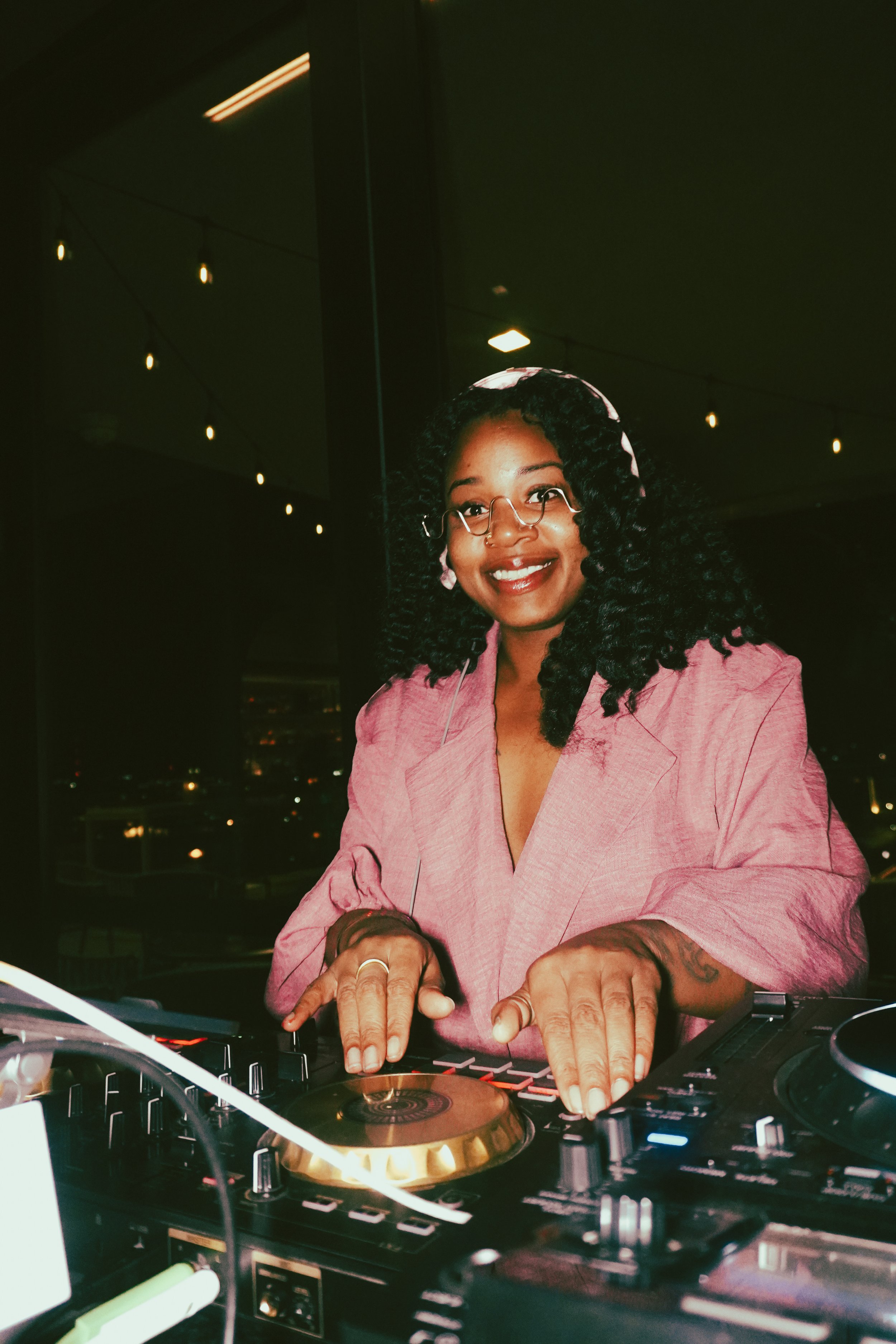 A woman with curly black hair, glasses, and a pink blazer, DJing at a nighttime event with city lights in the background.