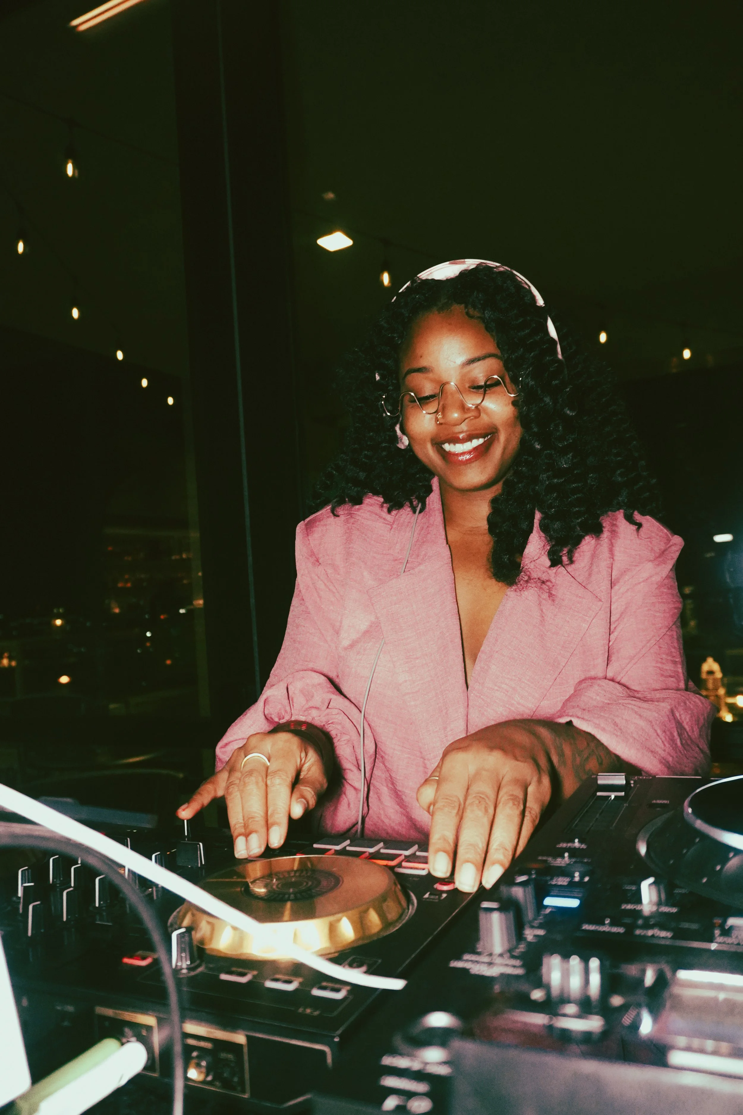 A woman DJ wearing glasses and a pink blazer, smiling and using DJ equipment at night.