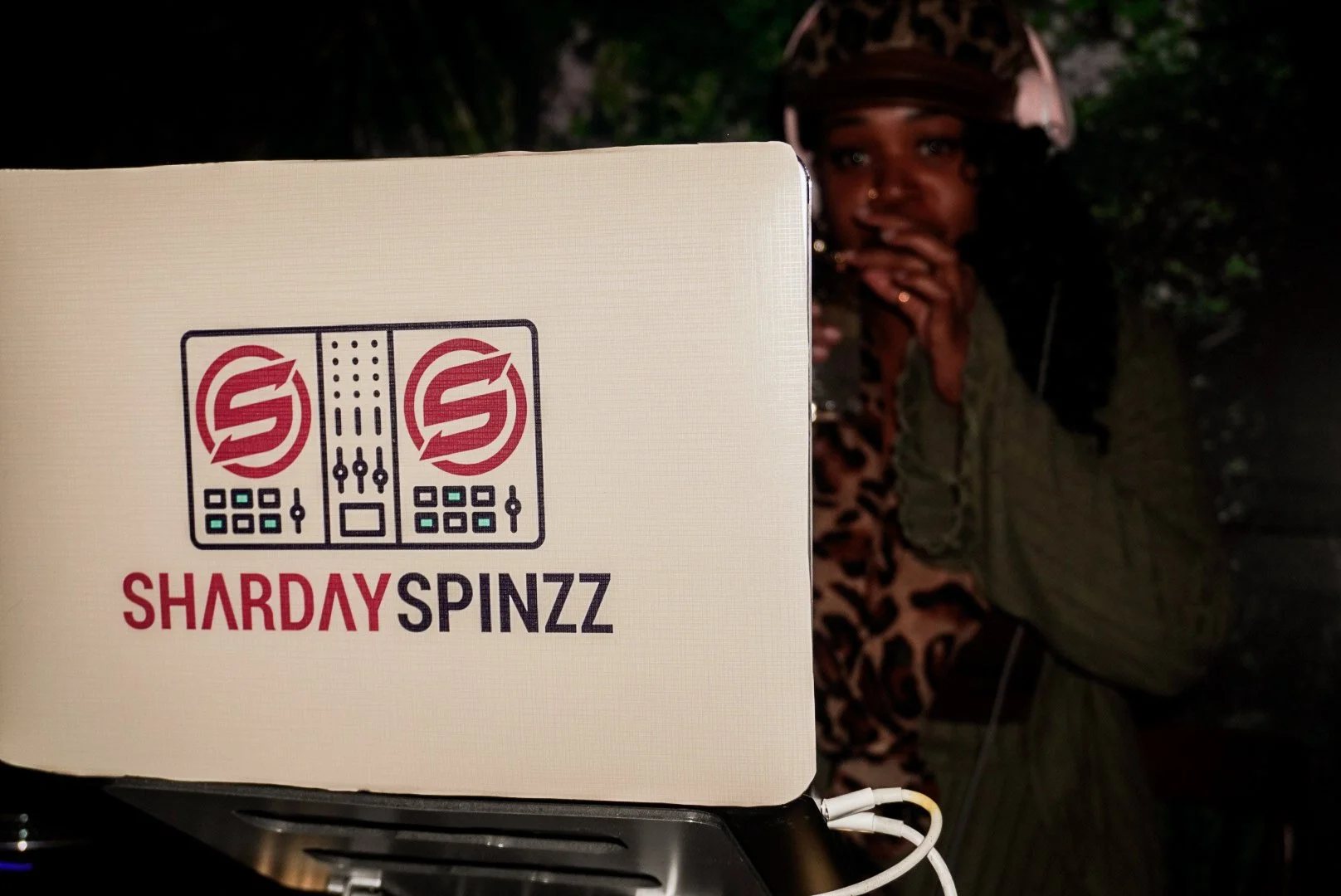 A person wearing a helmet and green jacket looking at the camera in the background, with a sign or box labeled 'SHARDAY SPINZZ' featuring a DJ graphic in the foreground.