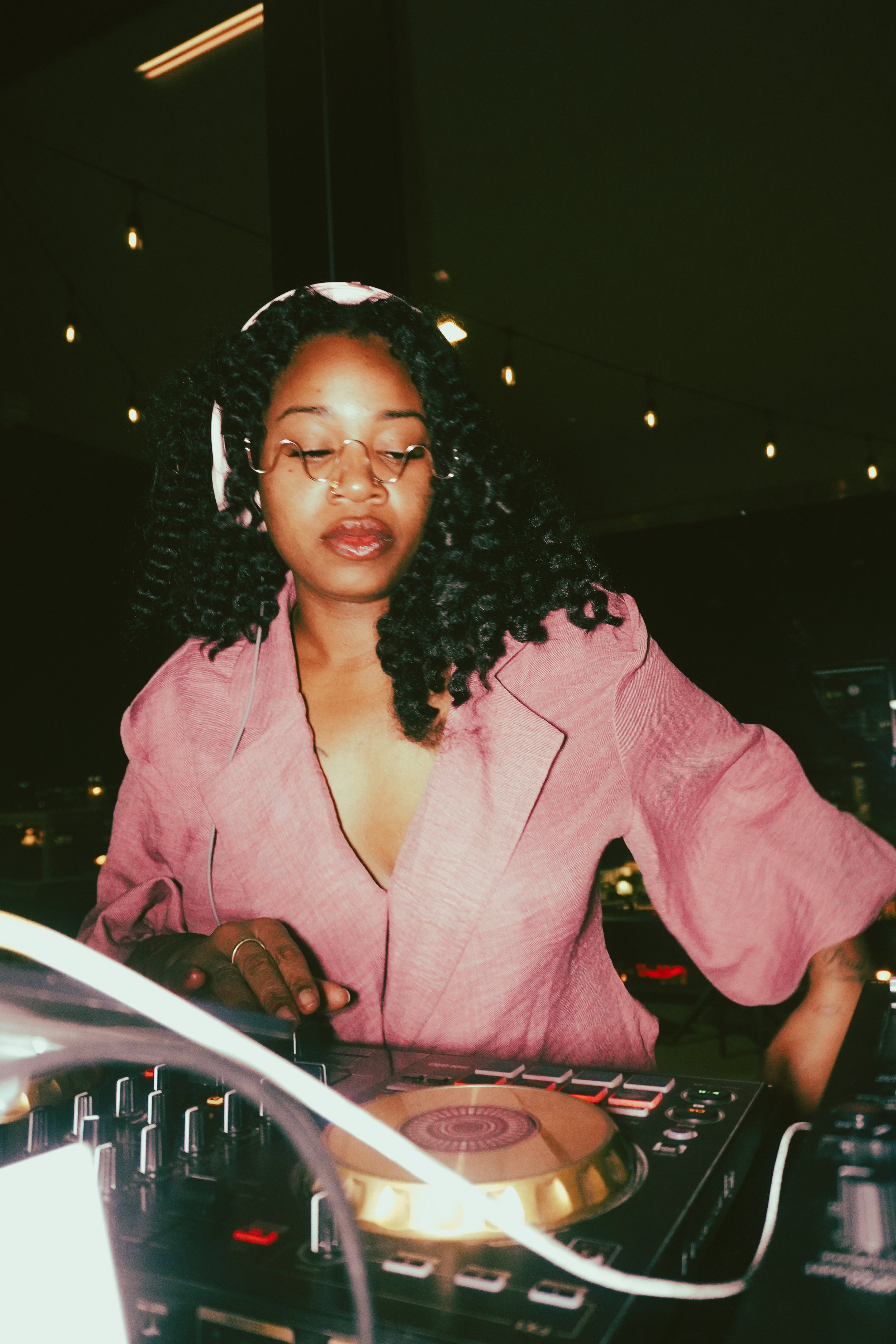 A woman with curly black hair, glasses, and a pink blazer standing at a DJ booth, adjusting musical equipment in a dimly lit environment with string lights overhead.
