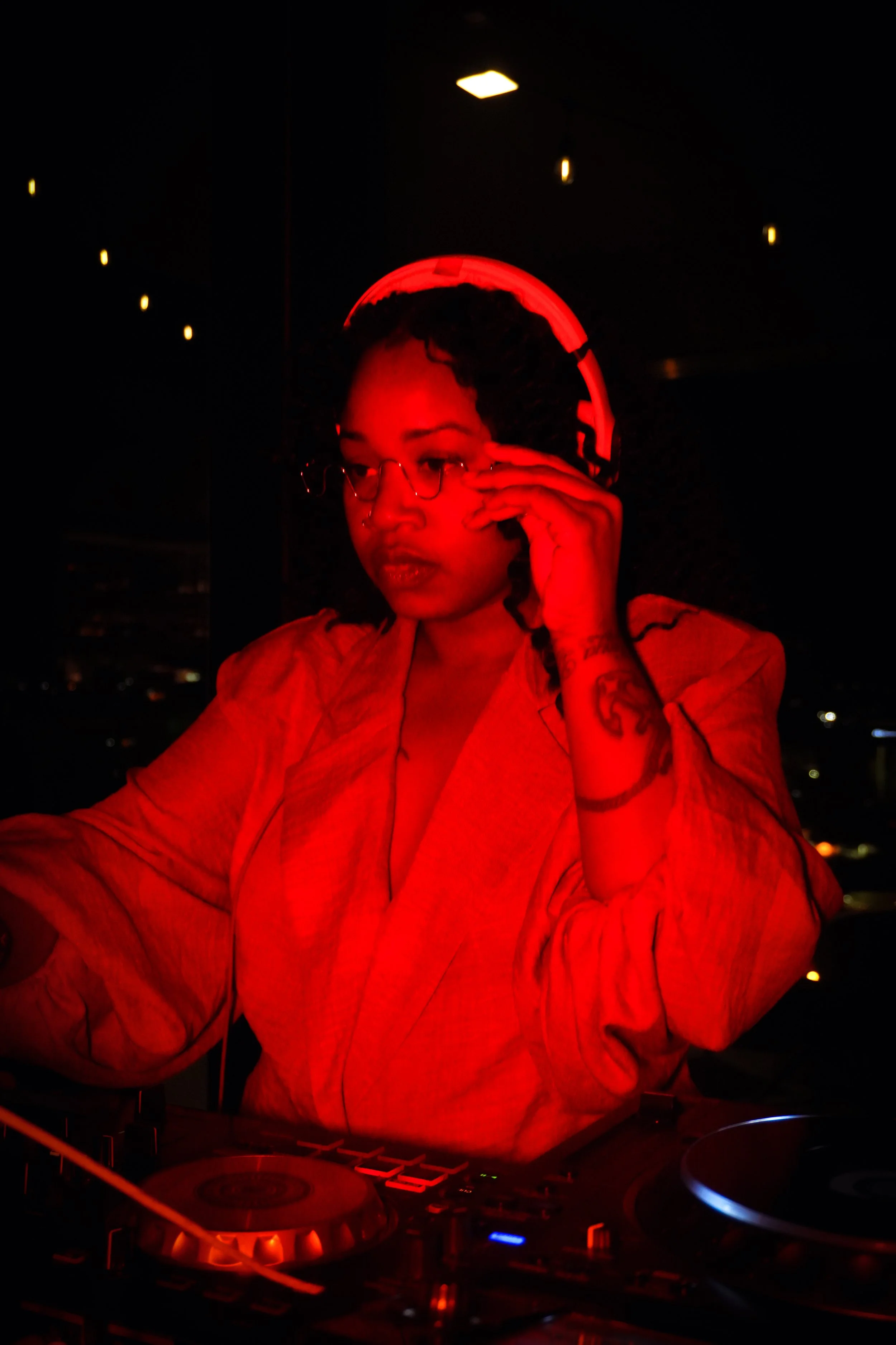 A woman with tattoos and glasses is DJing in a dark room, illuminated by red lighting.
