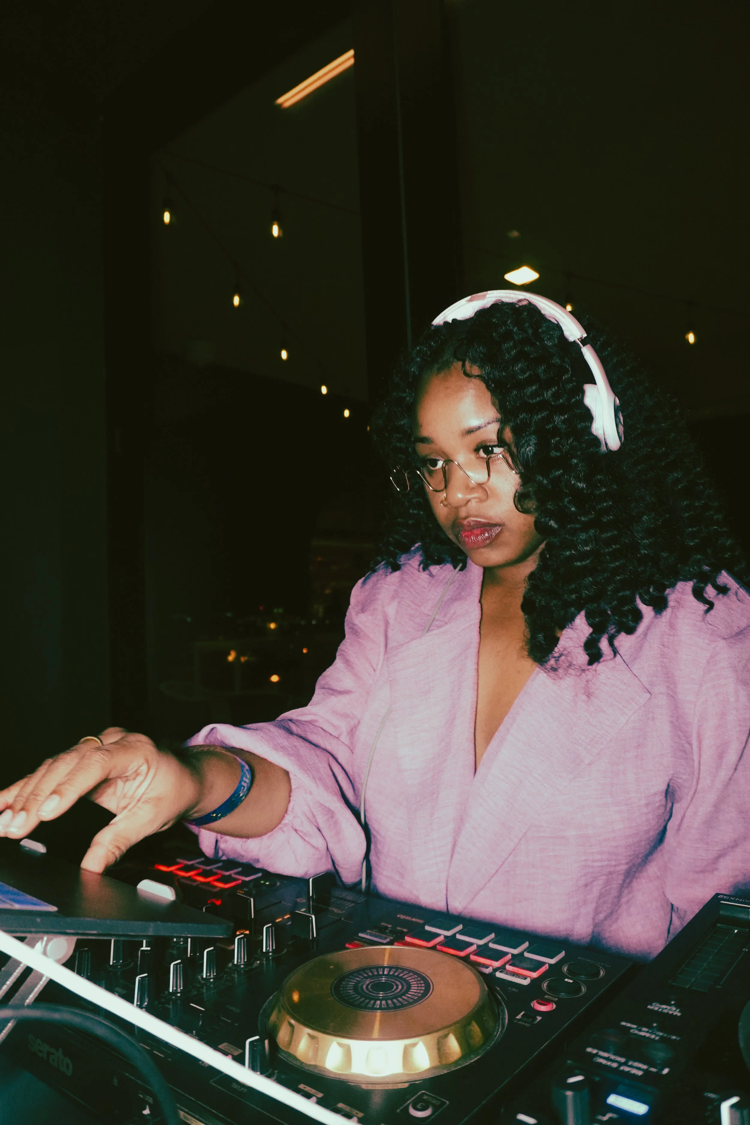 A female DJ with curly hair, glasses, and headphones, wearing a pink blazer, is operating a DJ mixer and turntable at a nighttime event with string lights in the background.