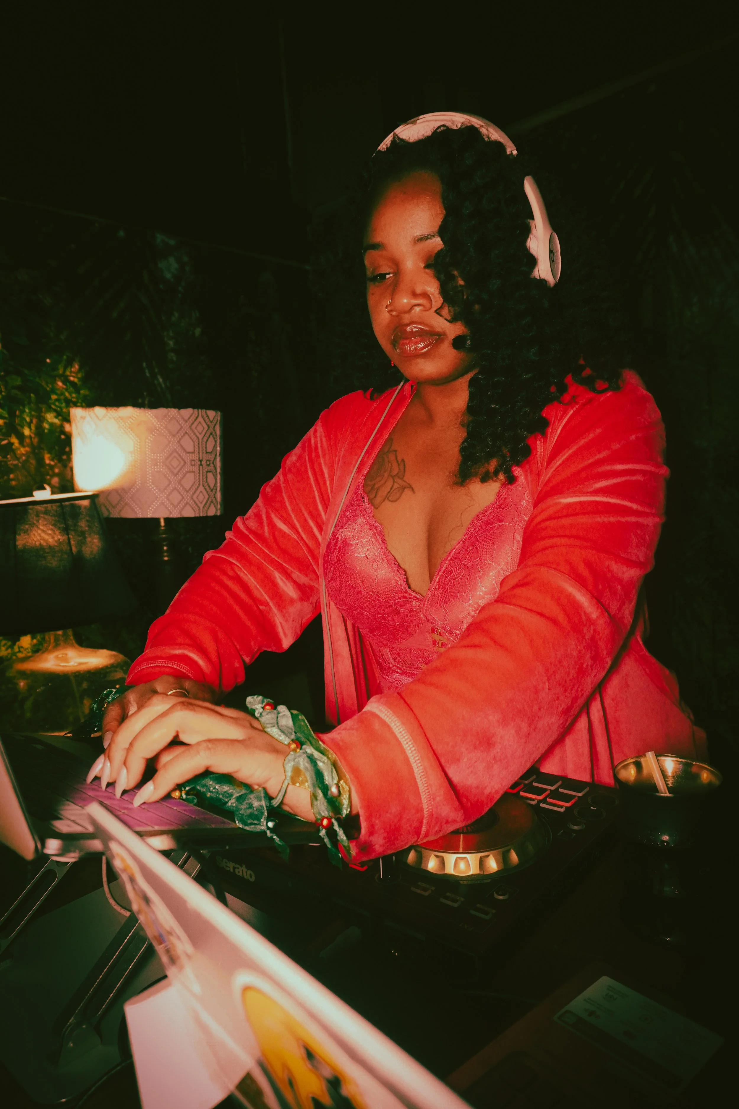 A woman with curly hair wearing a red outfit and headphones DJing at a dimly lit venue.