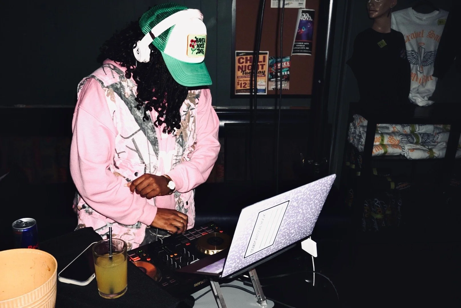 Person with long curly hair wearing a pink camouflage jacket, green and white trucker hat with 