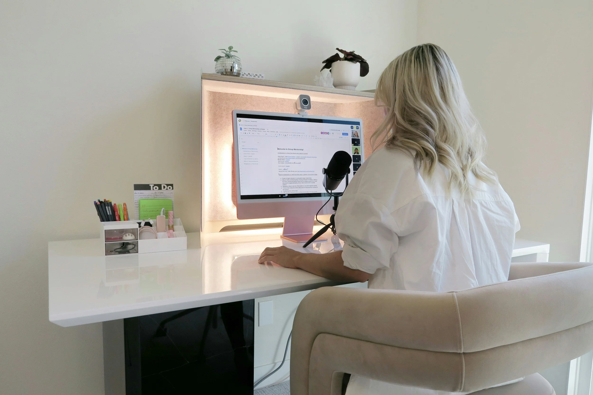 Woman with blonde hair using a computer at a white desk with a microphone, organizer, and notebook, in a bright room.