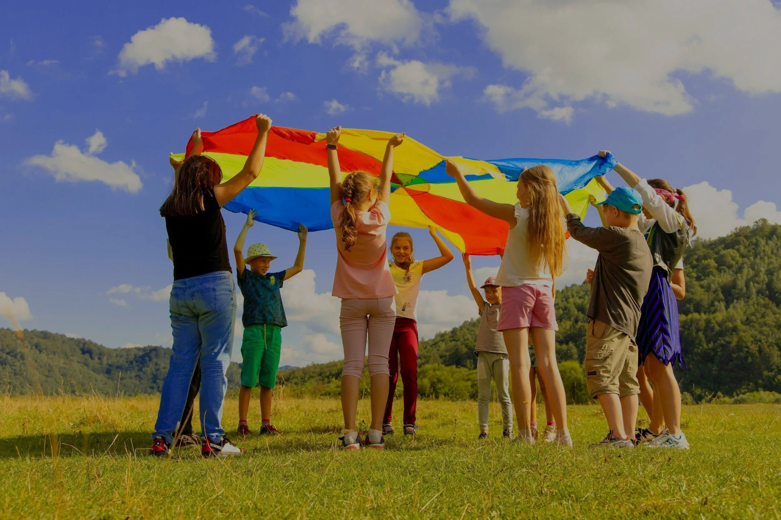 Children playing with a colorful parachute outdoors in a grassy field with hills and a blue sky in the background.