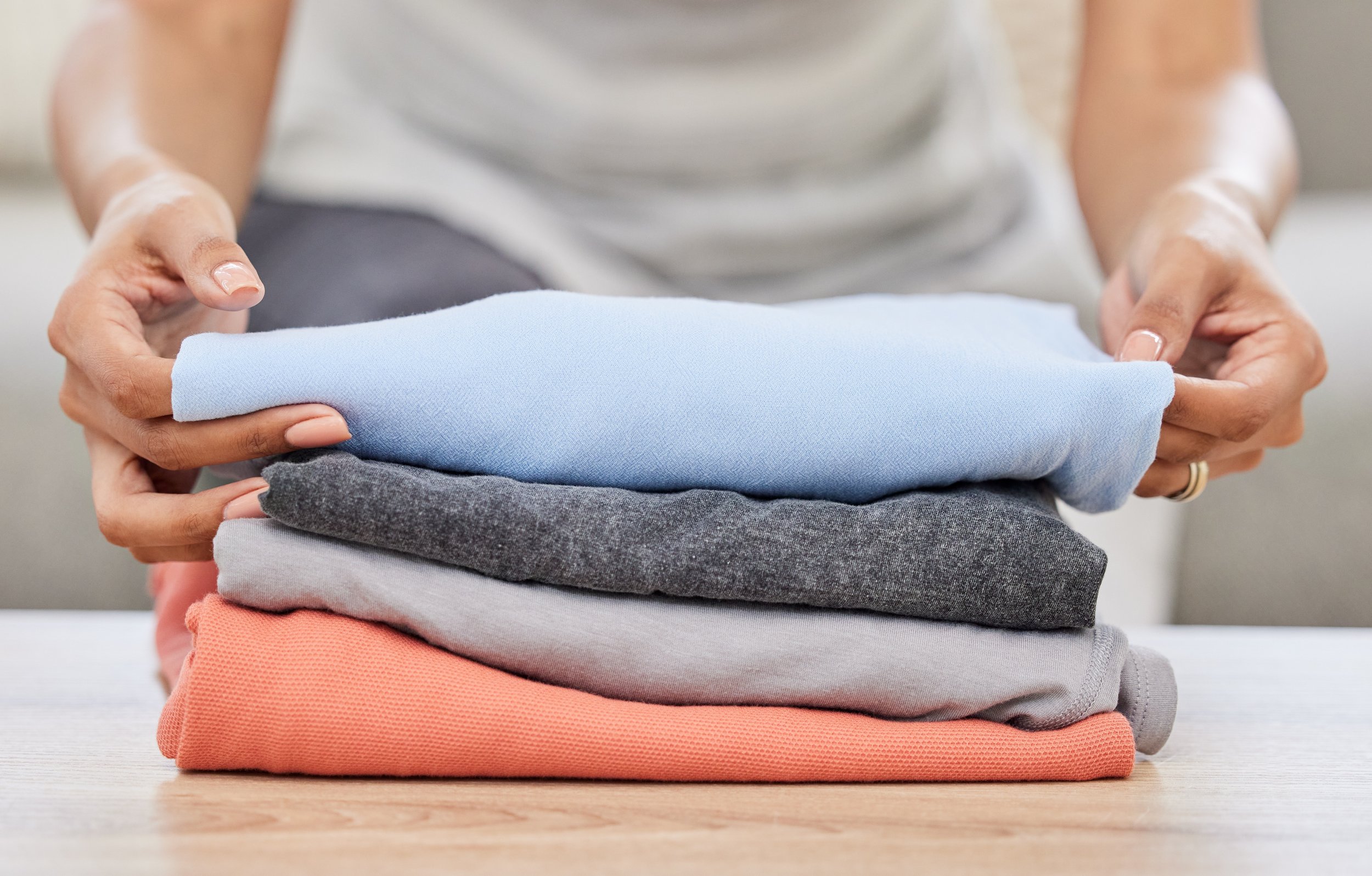 Person folding a stack of five folded shirts in different colors: blue, gray, left unspecified, coral, and beige, on a wooden surface.