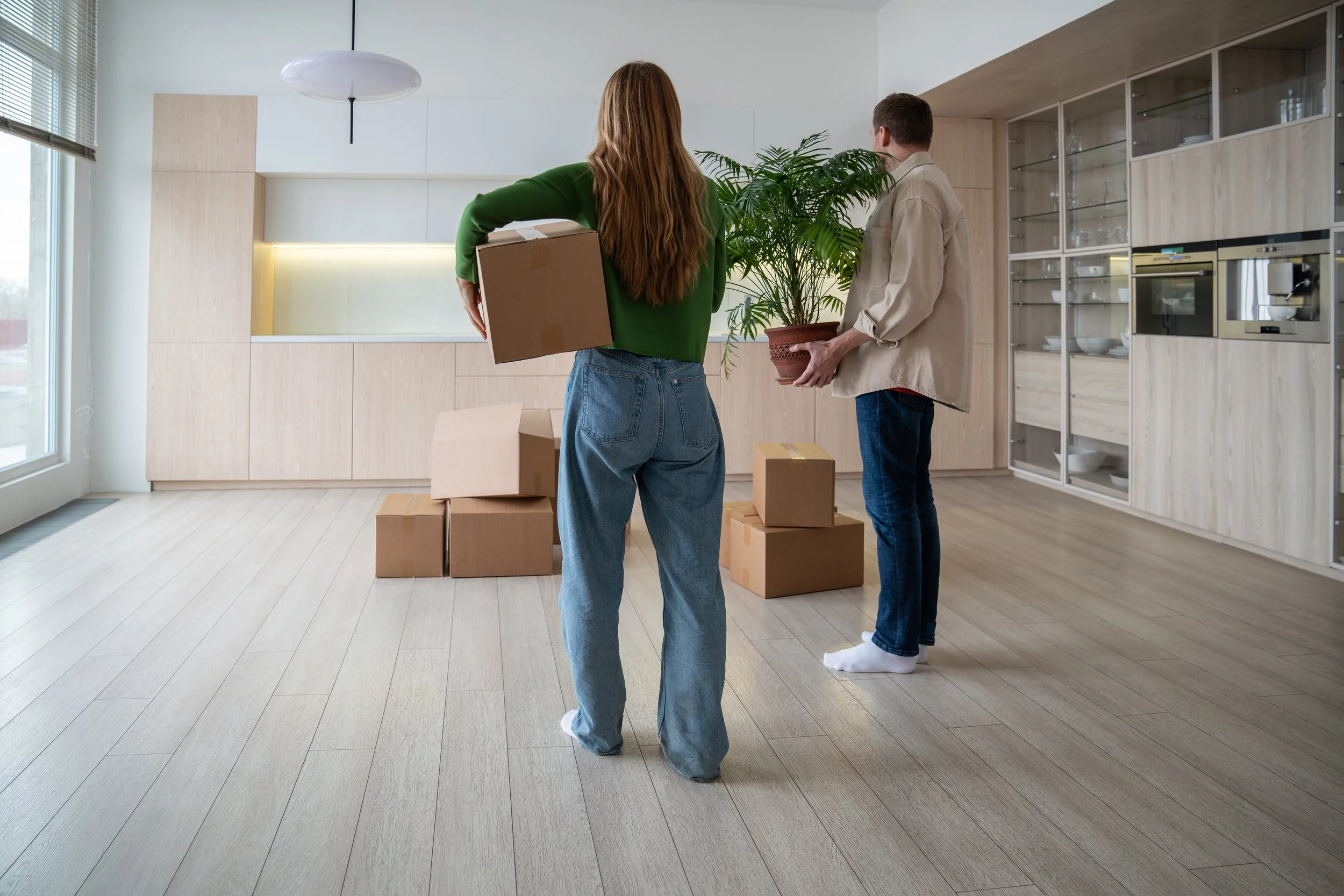 Two people moving into a new apartment, holding a potted plant and cardboard boxes, with more boxes on the floor.