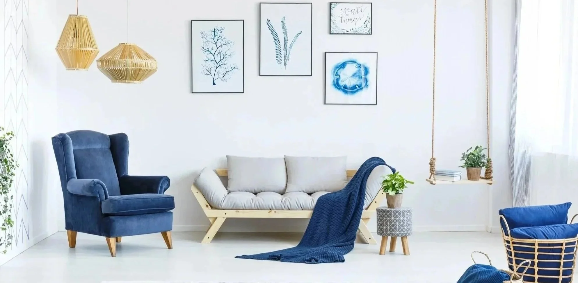 Living room with white walls, framed botanical artwork, hanging rattan lamps, and a swing shelf with books and plants. Furniture includes a blue armchair, a beige cushioned sofa with a blue throw blanket, and a small plant on a patterned side table.