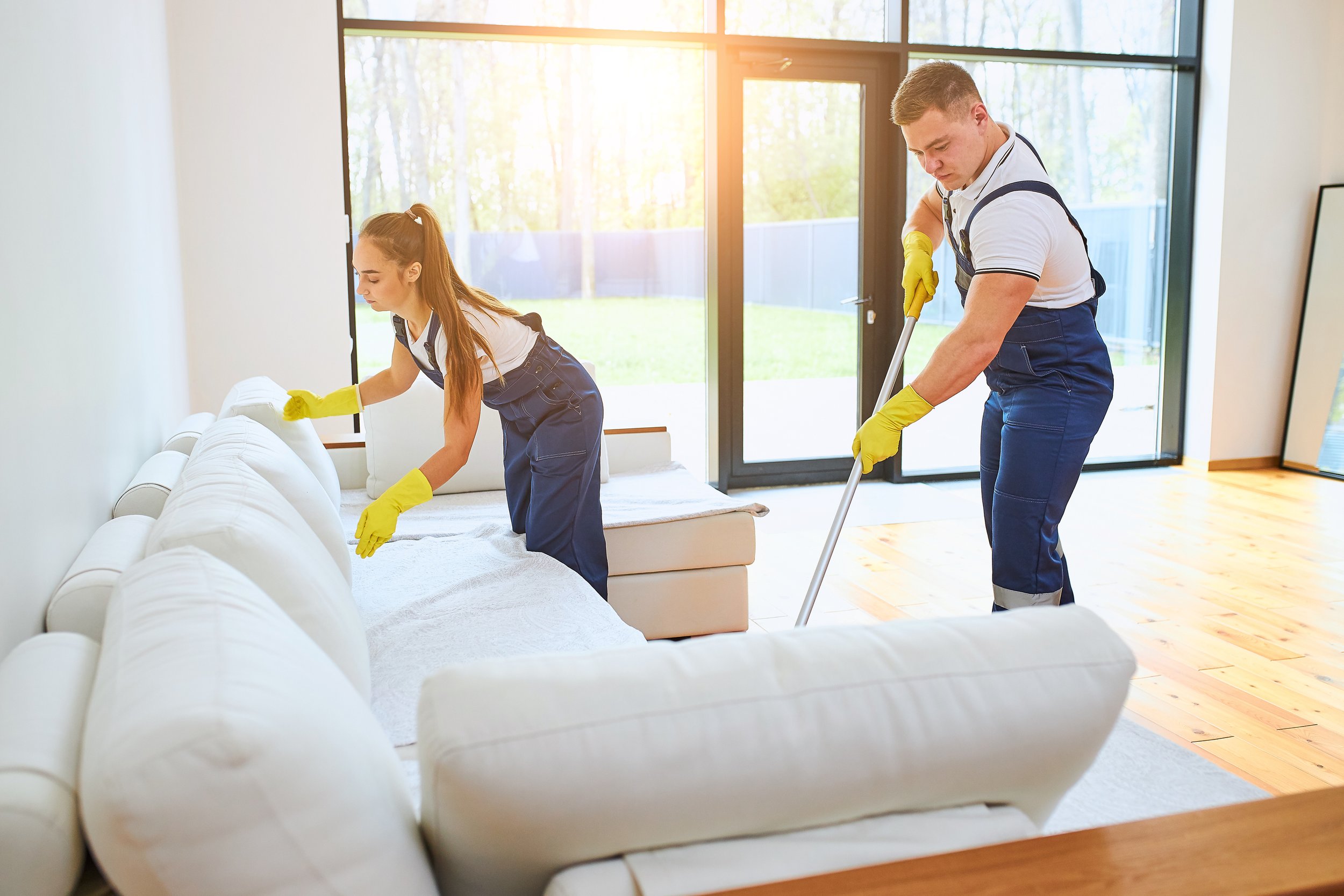 Two young adults, a man and a woman, cleaning a white sofa in a bright living room with large windows and wooden floors. They are wearing yellow cleaning gloves and casual clothes.