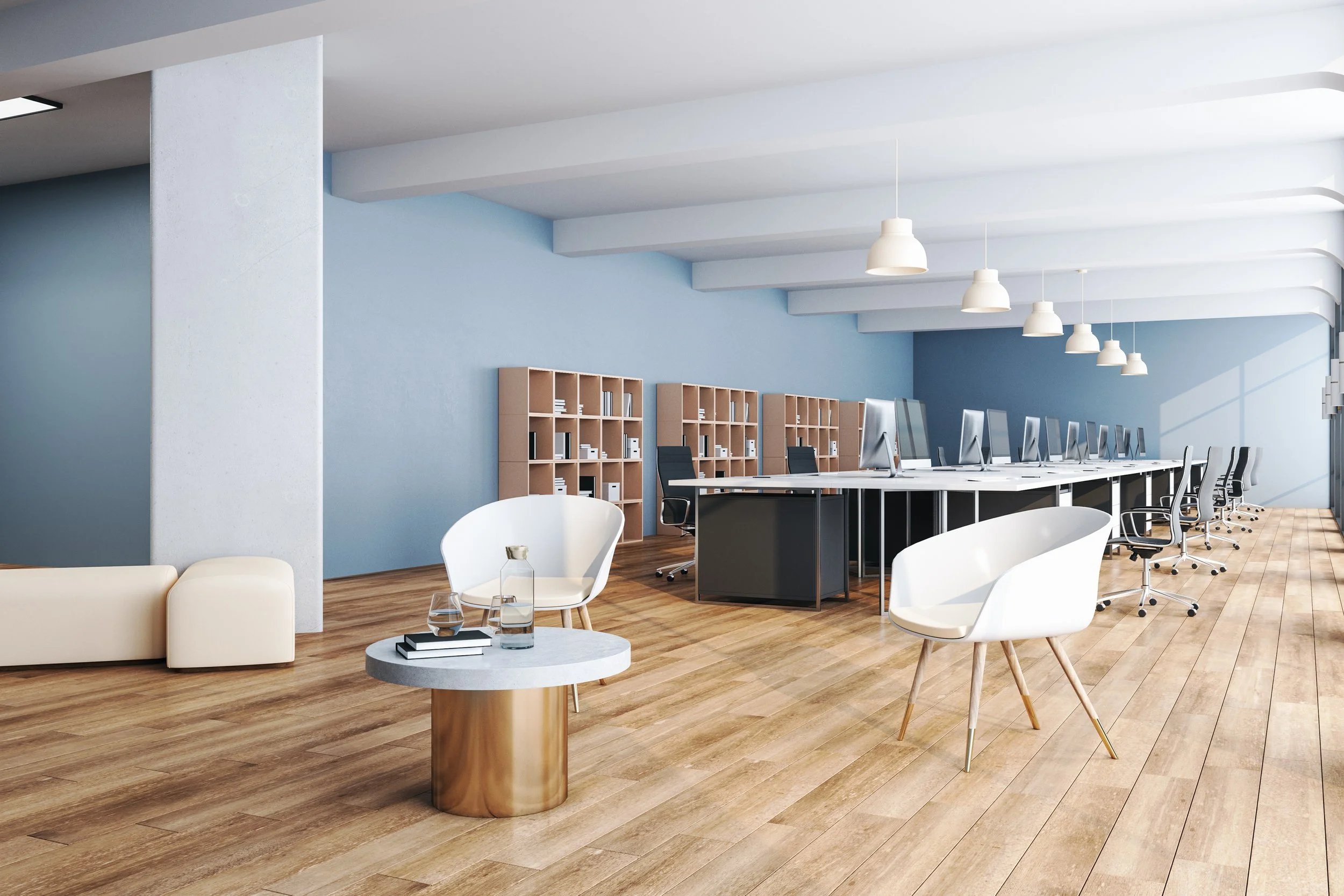 Modern office with a row of computers on a long table, blue walls, wooden flooring, bookshelves, white chairs, end table with glass items, and white ceiling lights.