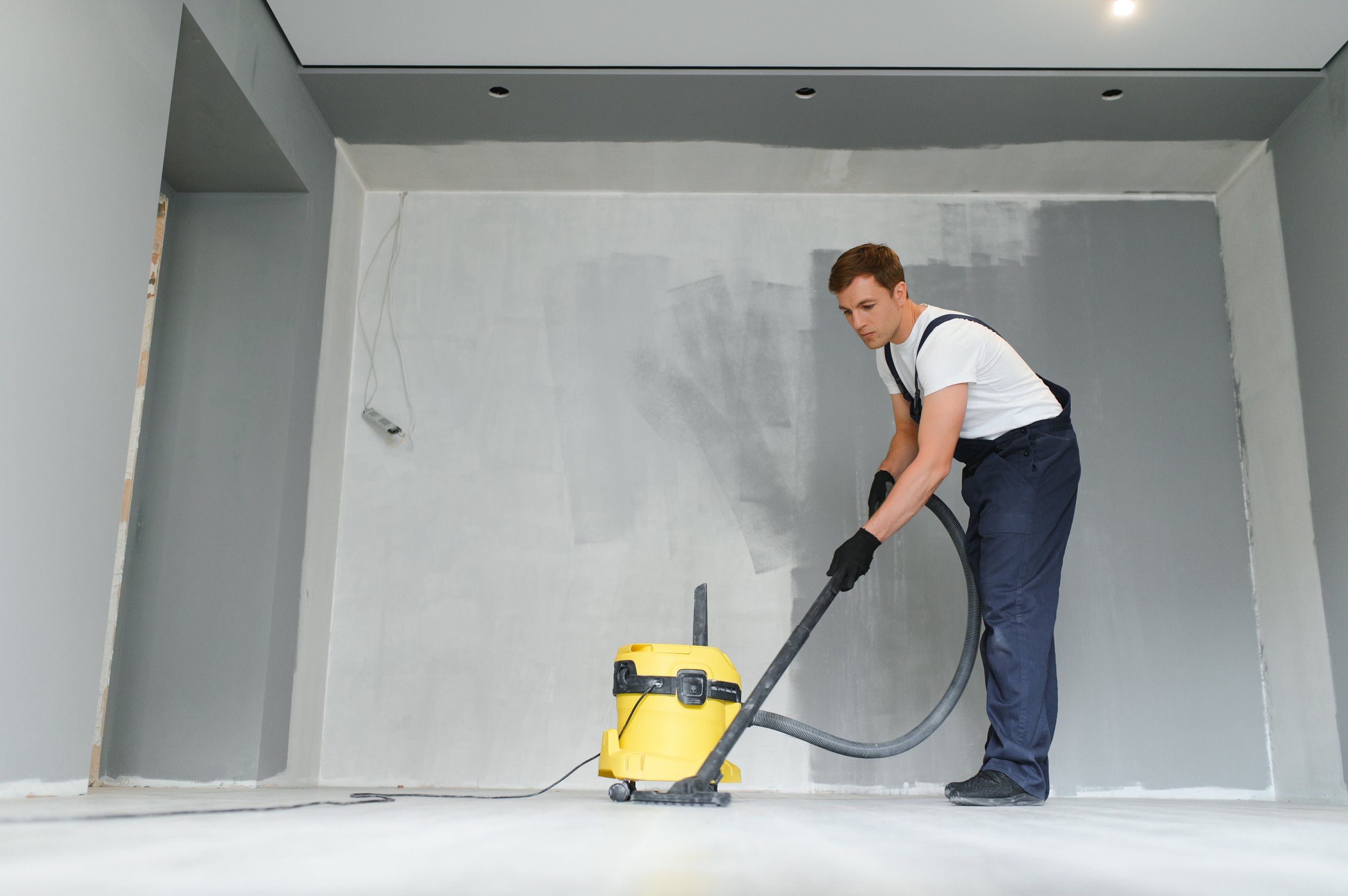 A man vacuuming a bare, unfinished room with gray walls.