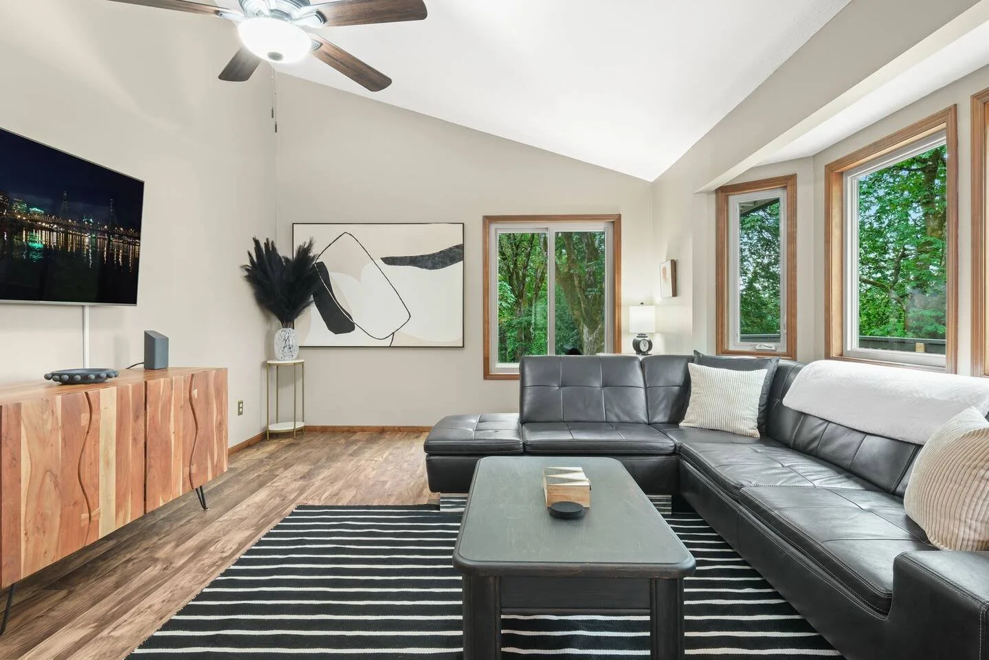 Living room with a black leather sectional sofa, a black and white striped rug, a wooden sideboard, a wall-mounted TV, and three large windows with greenery outside.