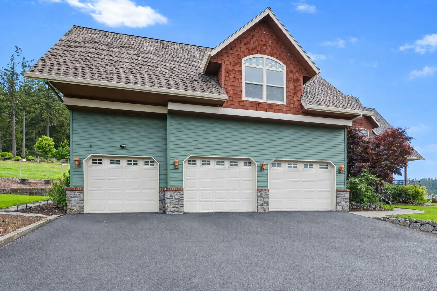 A house with a three-car garage, green siding, stone accents, and a brown shingle roof with a second-story window. The driveway is paved, and there is a landscaped yard with trees and plants.