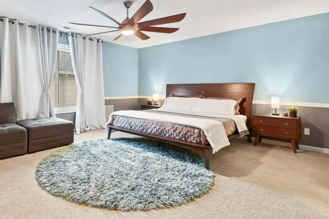 A bedroom with a bed, nightstands, lamps, a ceiling fan, a window with white curtains, a gray armchair, and a round shaggy rug.