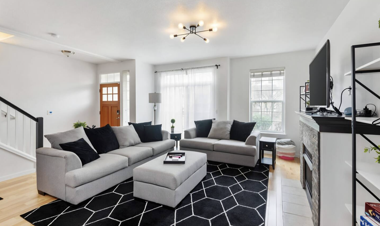 A bright living room with white walls and large windows, featuring a gray sectional sofa with black and gray pillows, a black and white geometric patterned rug, a television on a stand above a fireplace, and modern ceiling lighting.