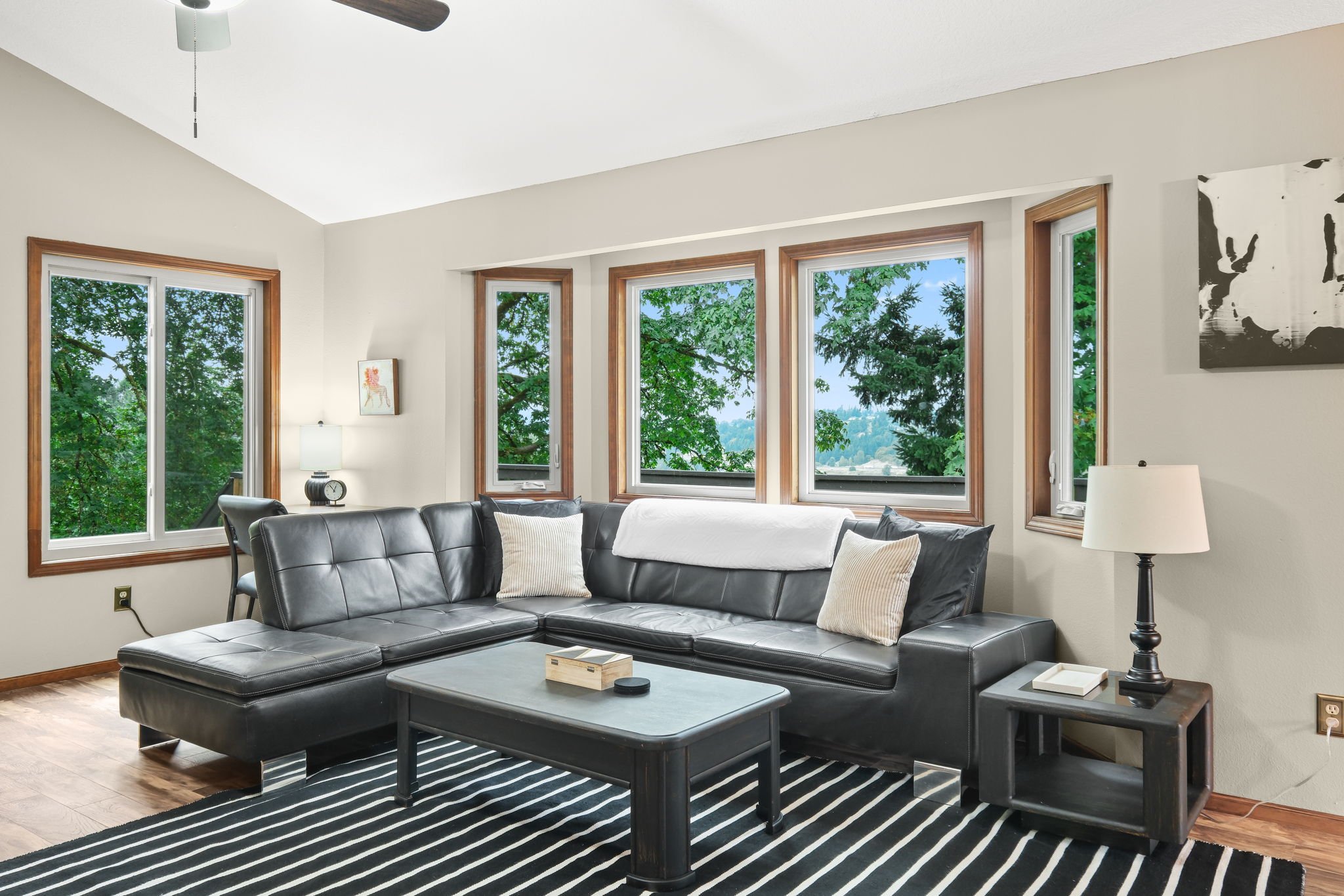 Living room with large windows showing green trees outside, a black leather sectional sofa with pillows, a black and white striped rug, a black coffee table, and side tables with lamps.