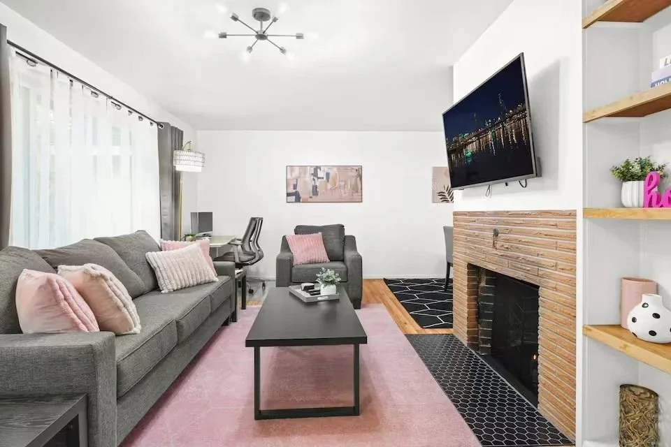 Modern living room with a gray sofa and loveseat, pink and neutral throw pillows, a black coffee table, a brick fireplace with a mounted TV, a pink rug, and a white bookshelf with decor.