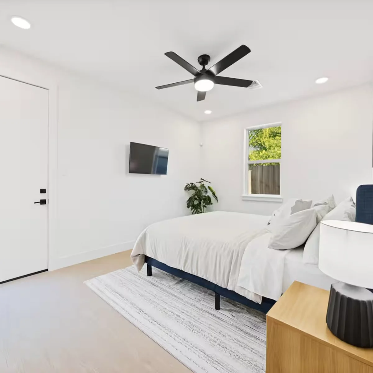 A modern bedroom with white walls, a bed with white linens, a wooden nightstand, a ceiling fan, a flat-screen television, a window with greenery outside, and a potted plant.