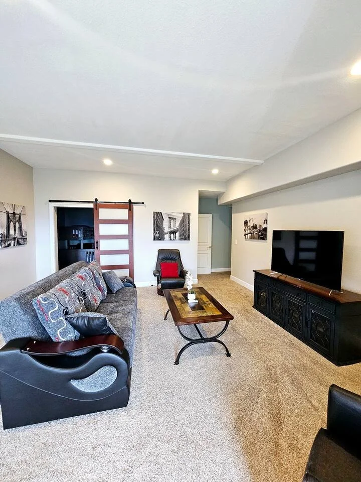Living room with a gray sofa, a black recliner with a red cushion, a wooden coffee table, a black TV stand with a large flat-screen TV, and black and white framed photographs on the walls.