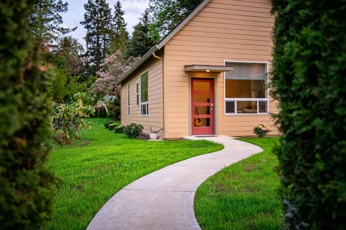 Front yard of a house with a winding concrete pathway leading to the front door, surrounded by green grass and shrubs.