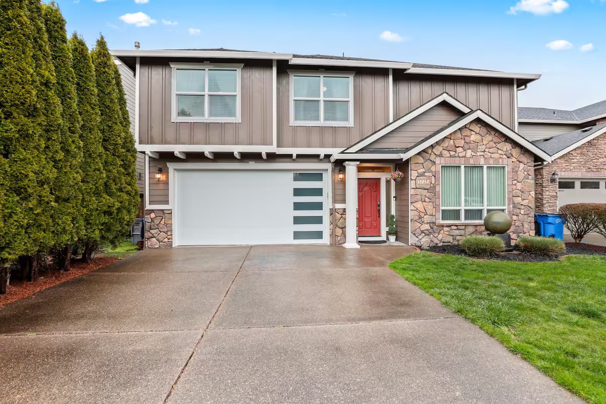 A two-story house with a stone and wood exterior, red front door, white garage door with horizontal glass panels, and well-maintained lawn and shrubs.