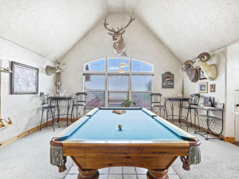 A room with a pool table in the center, decorated with mounted animal heads on the walls, large window with blinds, and small tables and chairs around the room.