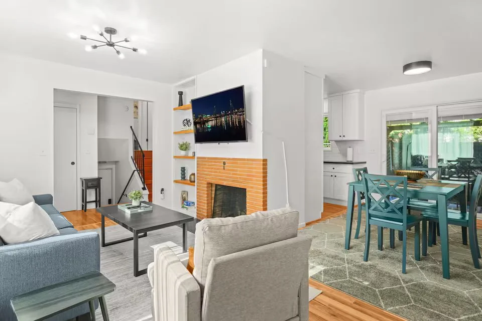 Living room with a gray sofa, beige armchair, black coffee table, and a wall-mounted flat-screen TV above a brick fireplace. Next to it, a dining table with six blue chairs and a green patterned rug. Open kitchen area with white cabinets and sliding glass door leading outside.