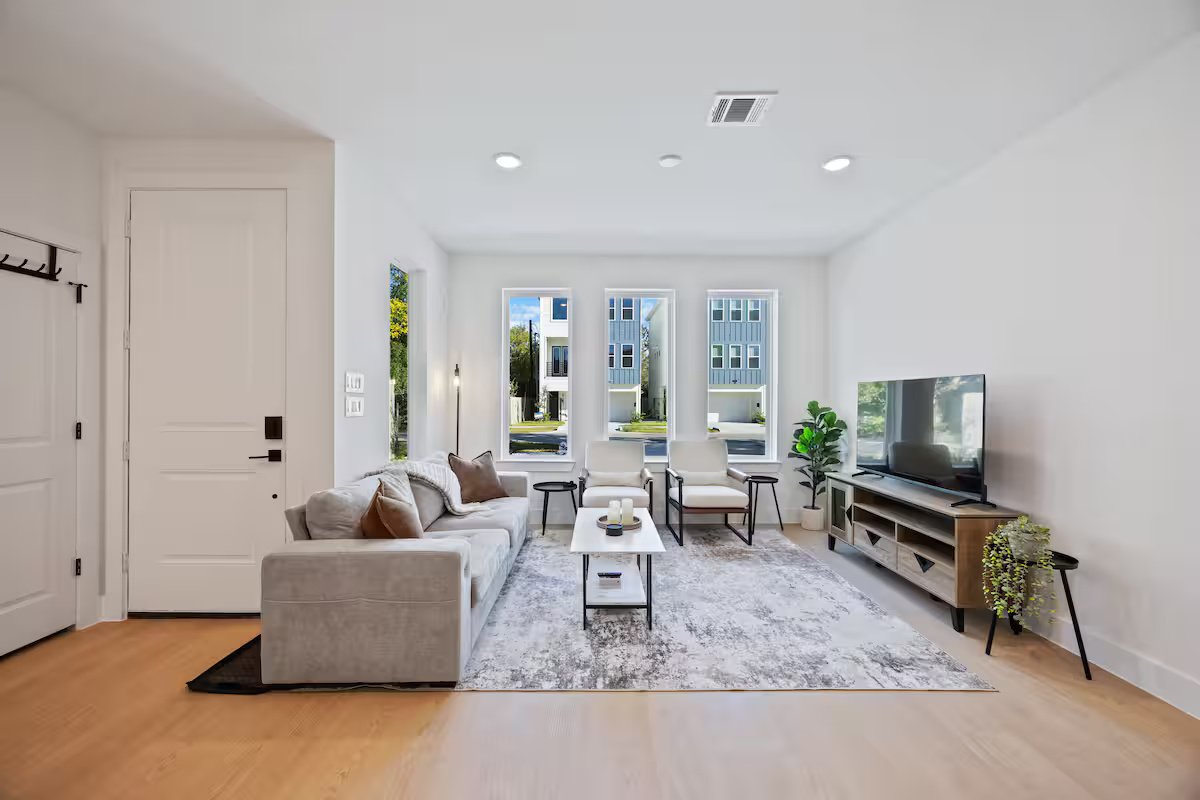 Bright, modern living room with white walls, large windows, a gray sofa with brown pillows, two white armchairs, a coffee table, a flat-screen TV on a wooden TV stand, indoor plants, a gray area rug, and natural light coming in from outside.