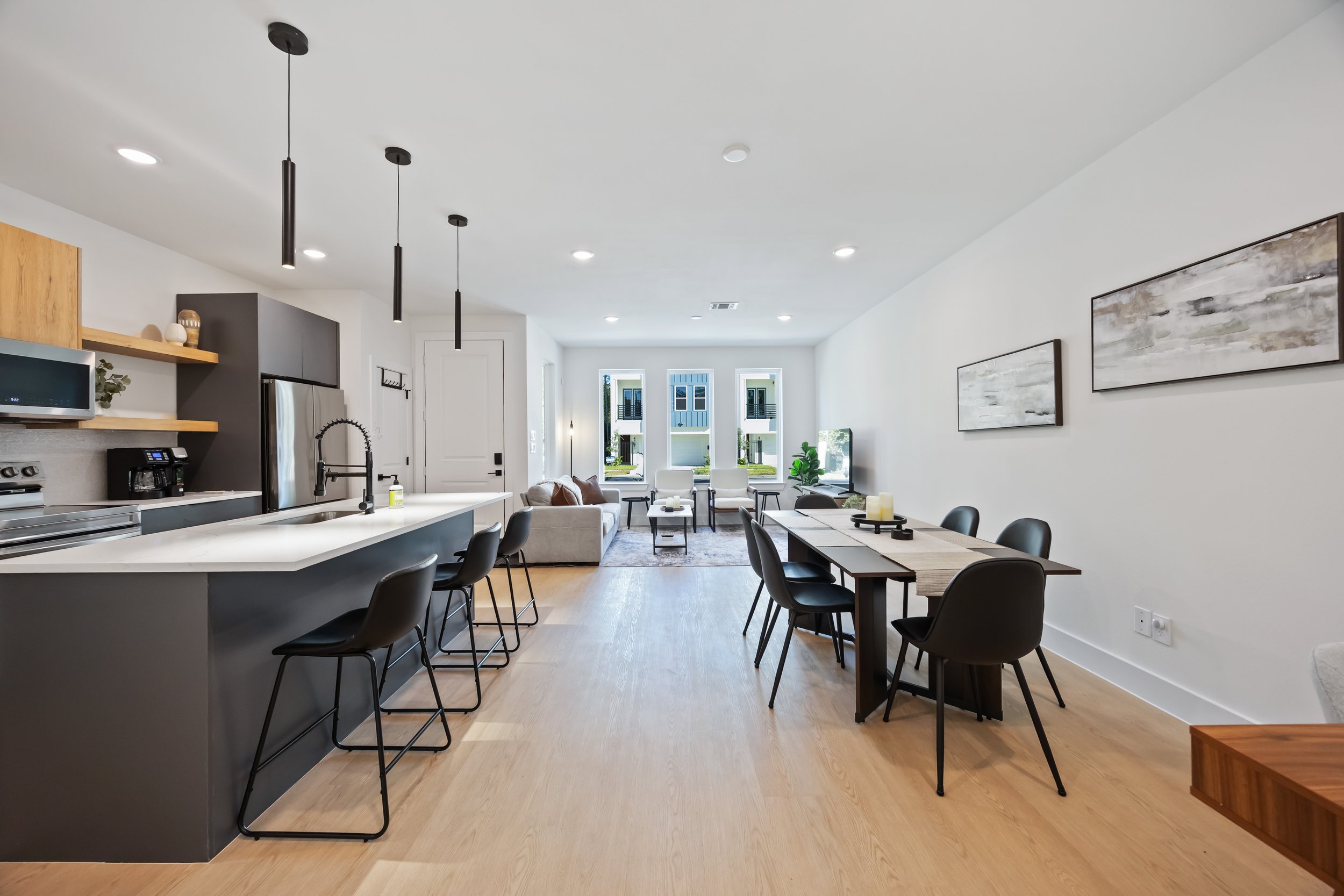 Open-concept living room and kitchen with modern decor, black dining chairs, a white island with black stools, dining table with black chairs, light wood flooring, and a large window with a view of the outside.