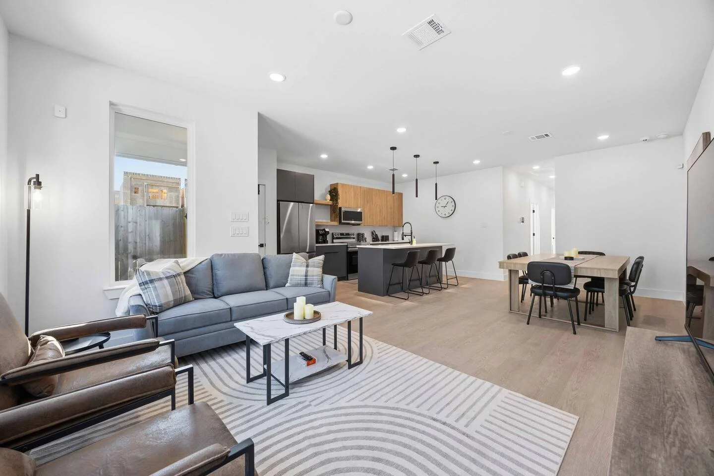 Open-concept living room and kitchen with modern furniture, wooden accents, and ample lighting.