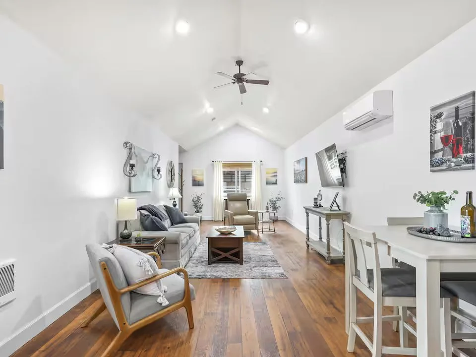 Living room with white walls, wood flooring, gray sofa, white armchair, coffee table, dining table with chairs, mounted TV, ceiling fan, air conditioning unit, decorative artwork, and plants.