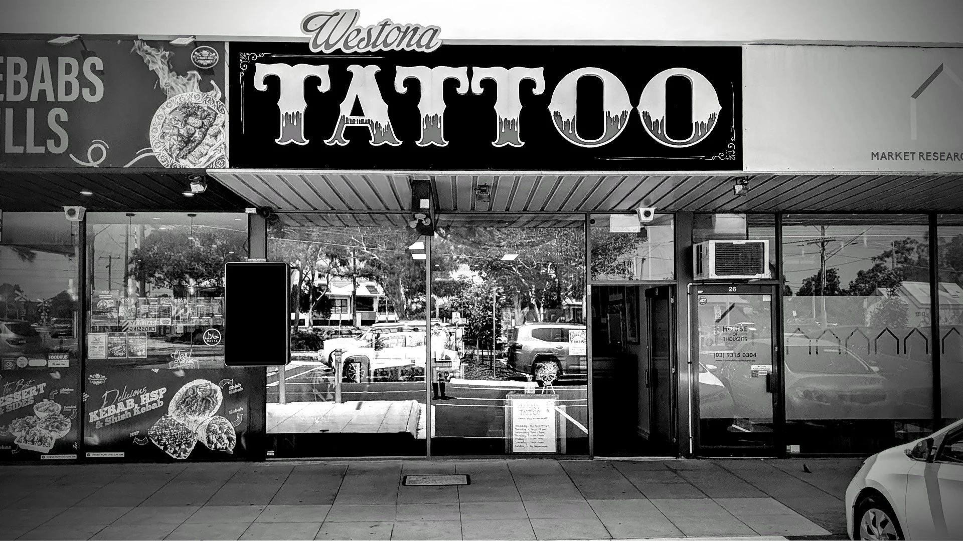 Exterior of a tattoo shop with large signage reading 'Westona TATTOO' and promotional posters on the windows advertising kebabs and other food items.