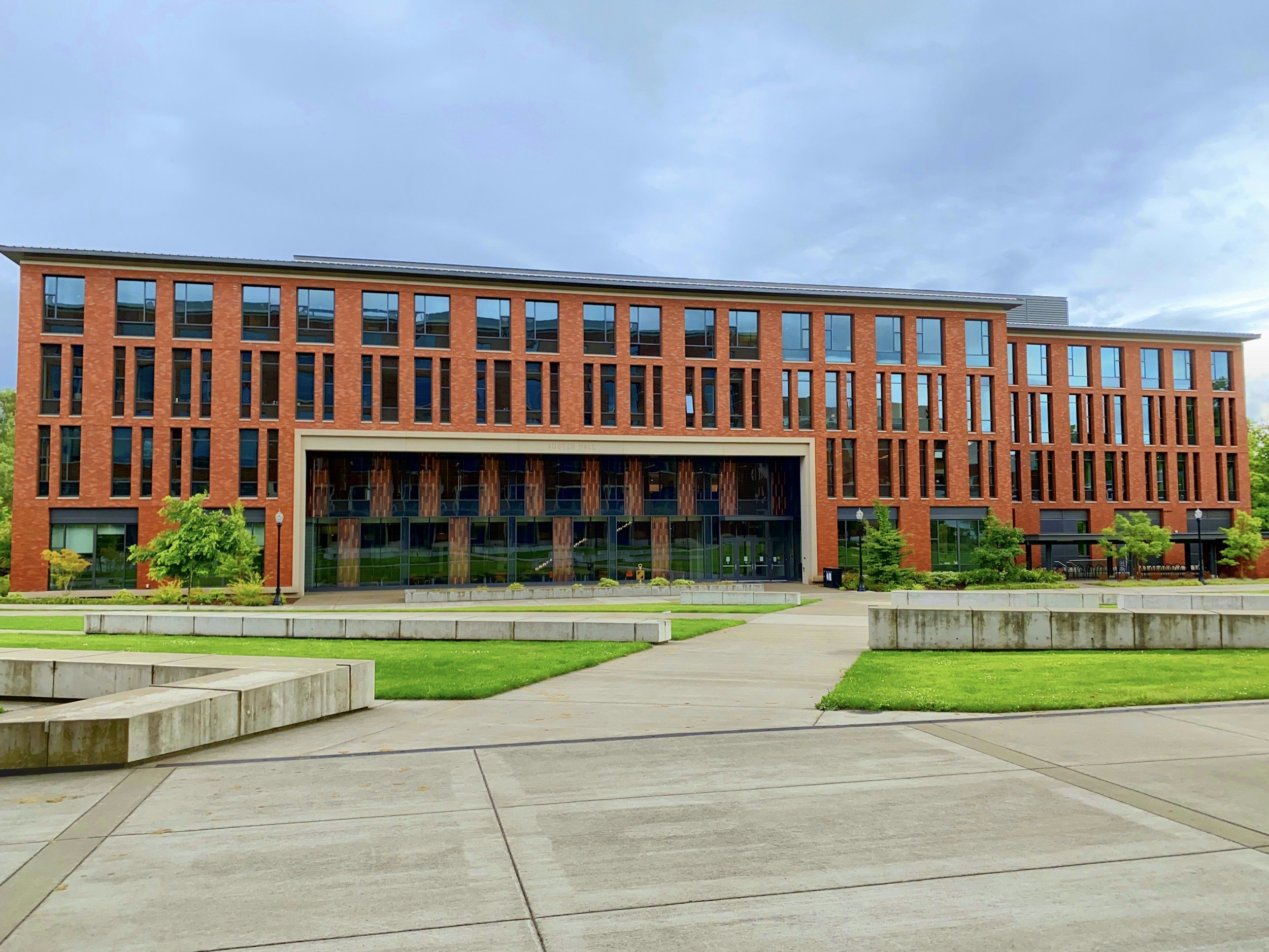 A modern multi-story building with a red brick facade, large windows, and a flat roof, surrounded by a landscaped area with trees and a concrete walkway.