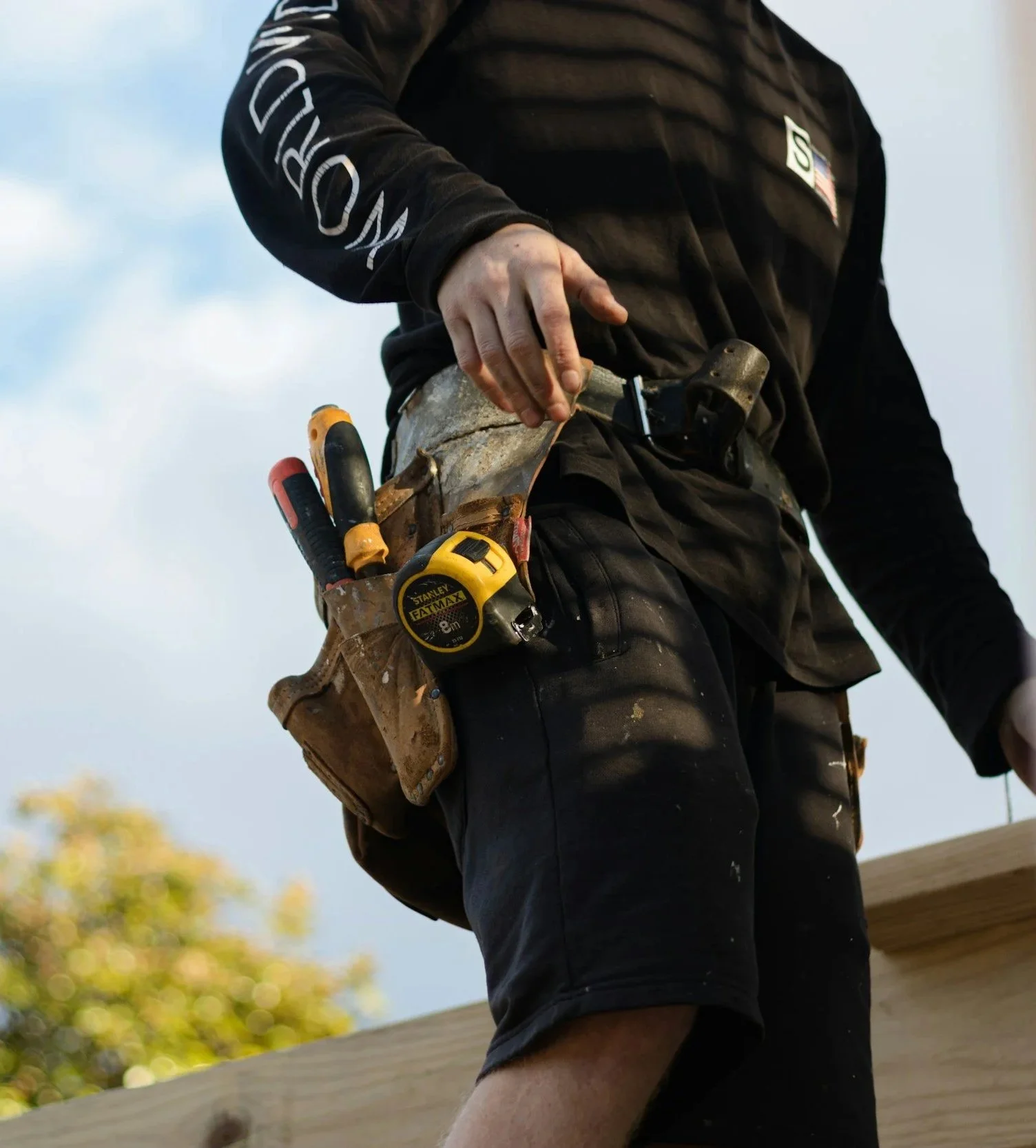 Close-up of a construction worker's waist, showing a leather tool belt with tools including a tape measure and screwdrivers, and part of their black long-sleeve shirt and shorts, working outdoors.