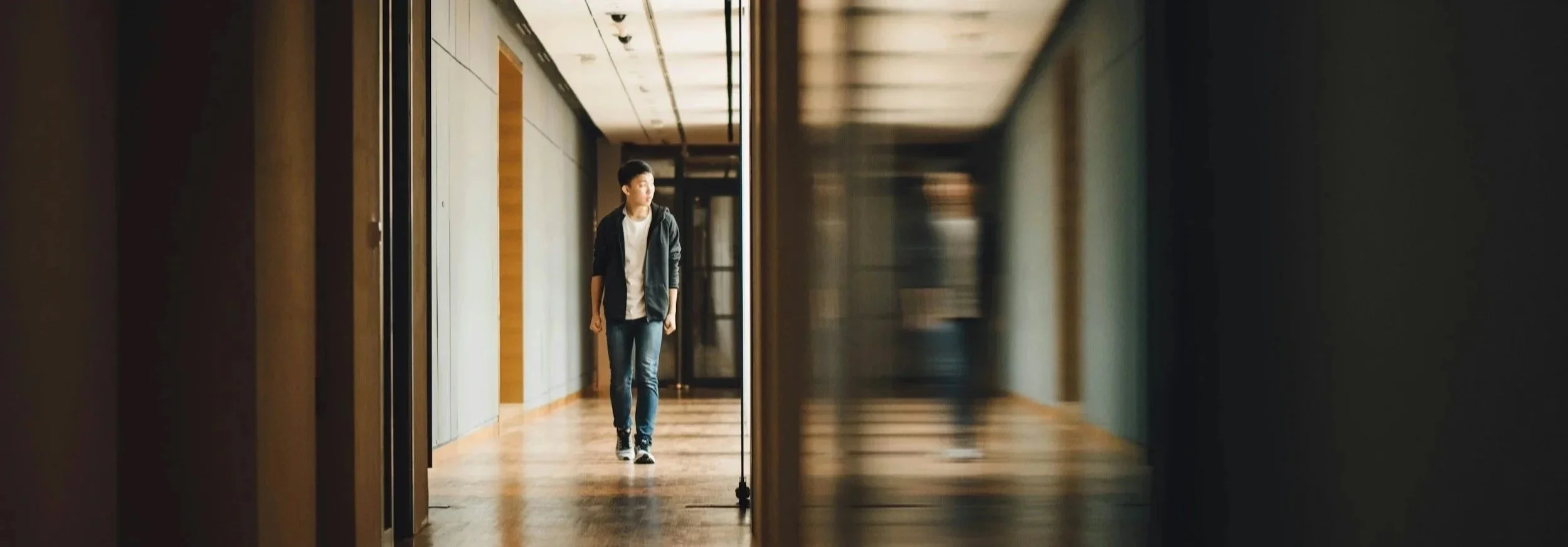 A young man walking down a hallway in a modern building with wooden floors and glass walls.