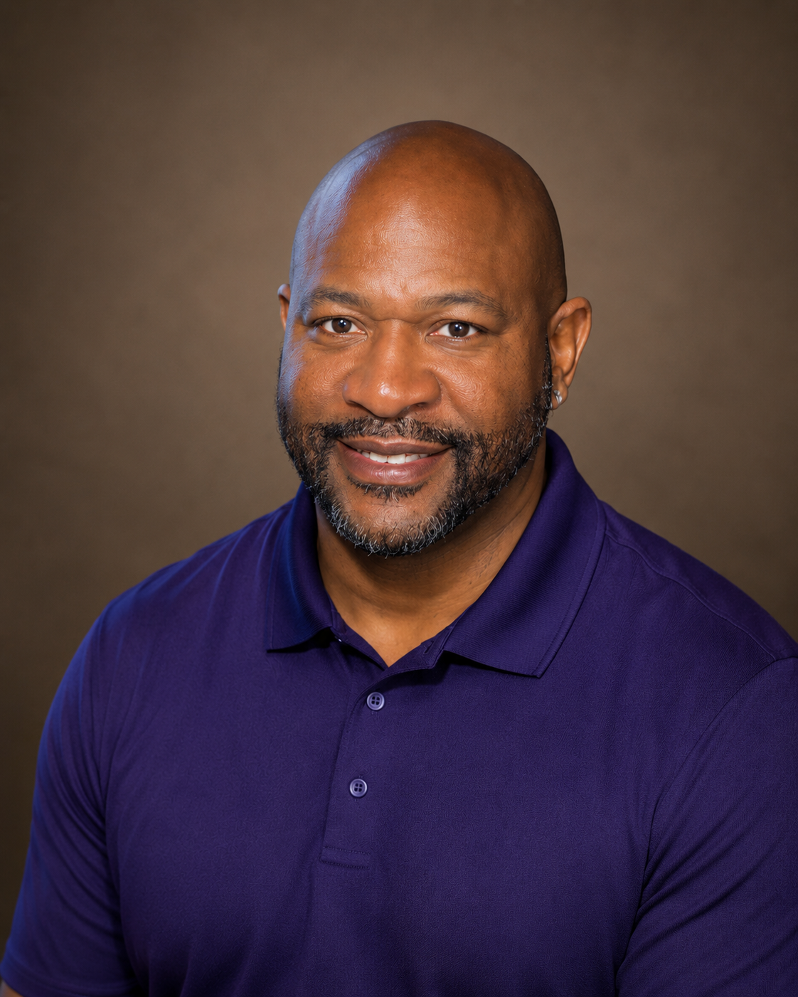 Portrait of a smiling African American man with a beard, wearing a purple polo shirt, against a brown gradient background.