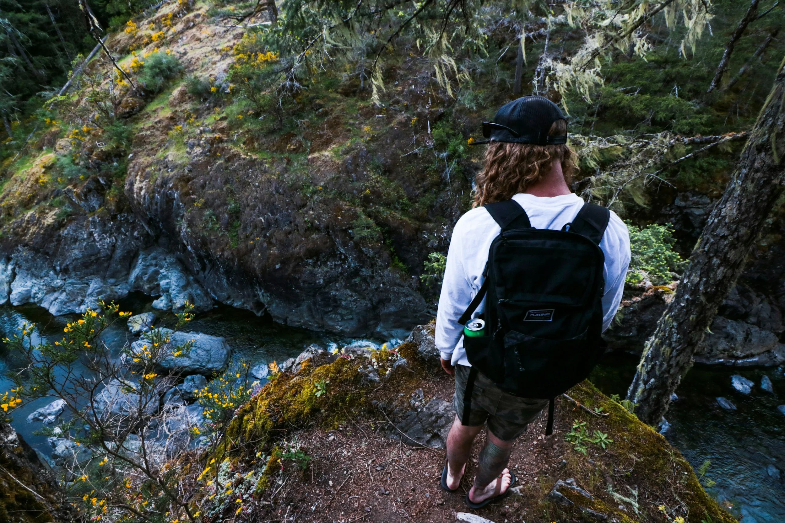 A person with long curly hair wearing a black cap, white long-sleeved shirt, camouflage shorts, and flip-flops, standing on a trail above a rocky stream in a forest.