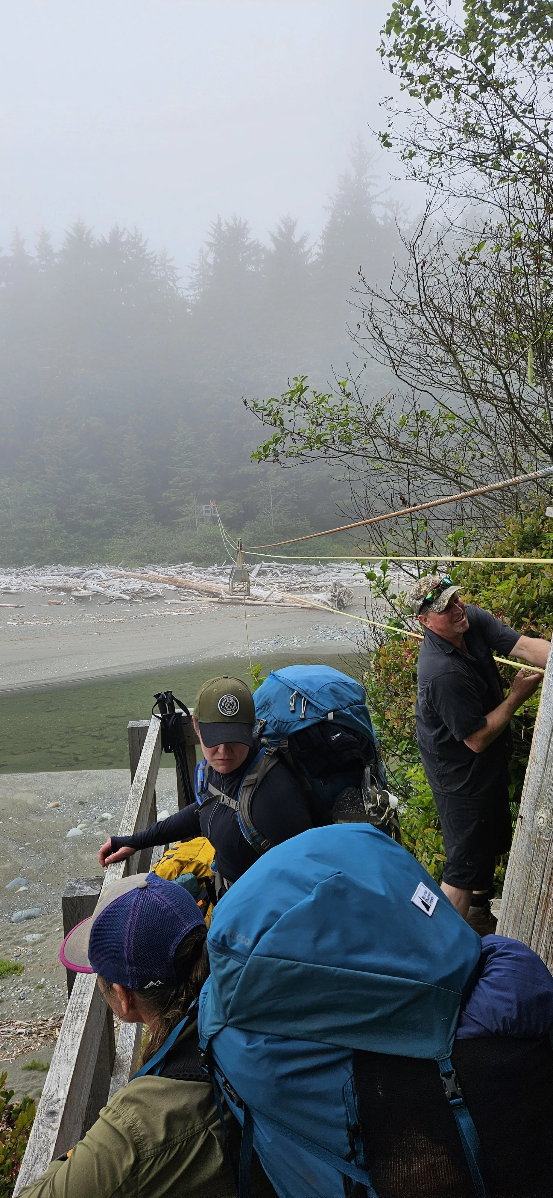 Three people with backpacks and outdoor clothing climbing on a wooden structure near a foggy river, surrounded by trees.