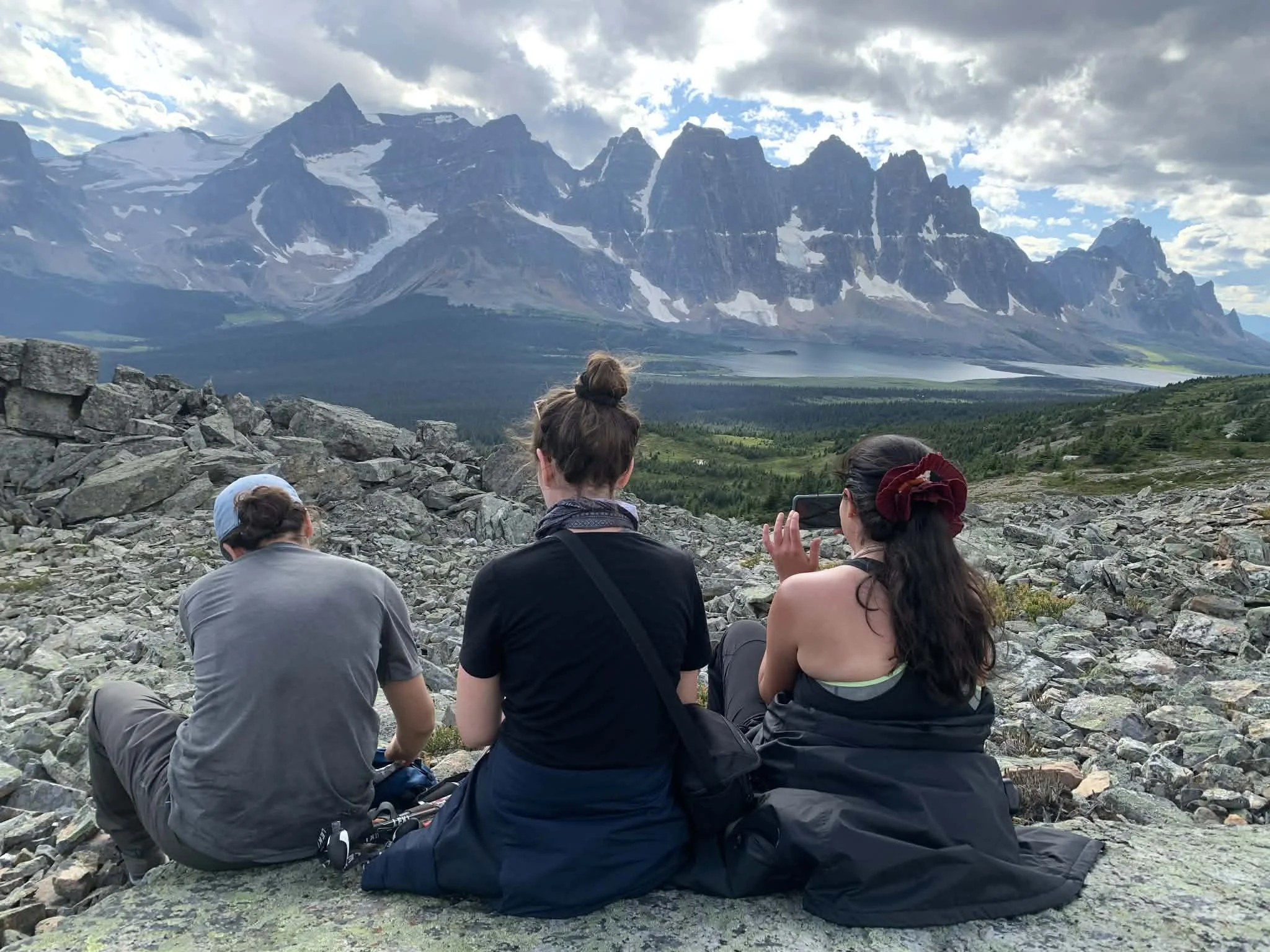 Three women and one man sitting on rocks, facing a mountain range with snow-capped peaks and a partly cloudy sky.