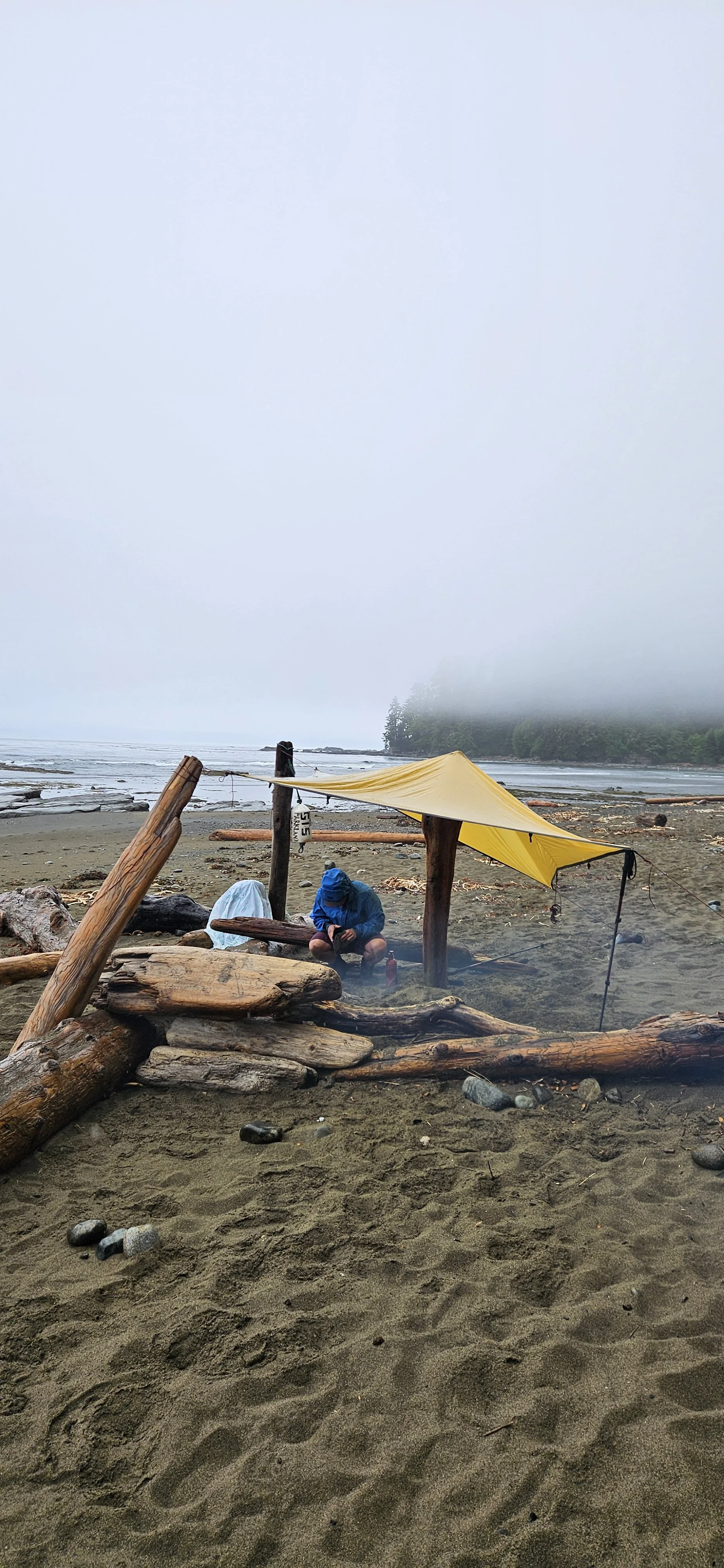 Person sitting under a yellow tarp on a sandy beach with driftwood and rocks, overlooking a foggy shoreline with water and trees in the background.
