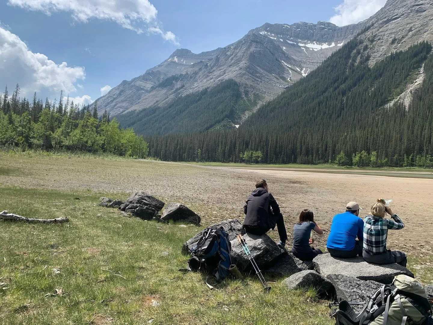 Four people sitting on rocks near a grassy area and a dry lakebed, with mountains, pine trees, and a partly cloudy sky in the background.