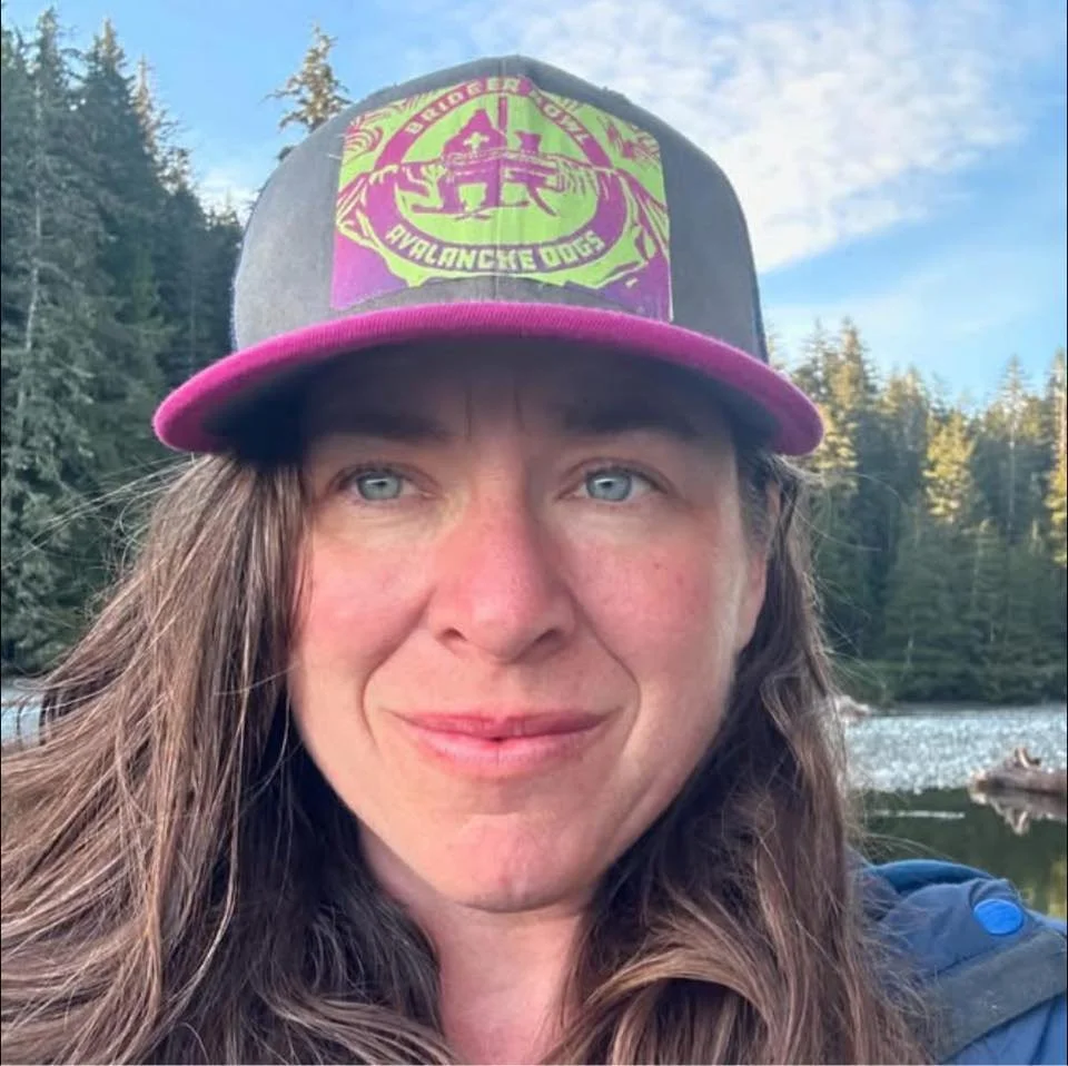 Close-up of a woman with long brown hair and blue eyes, wearing a black cap with a green and purple patch, outdoors in a forested area near water.