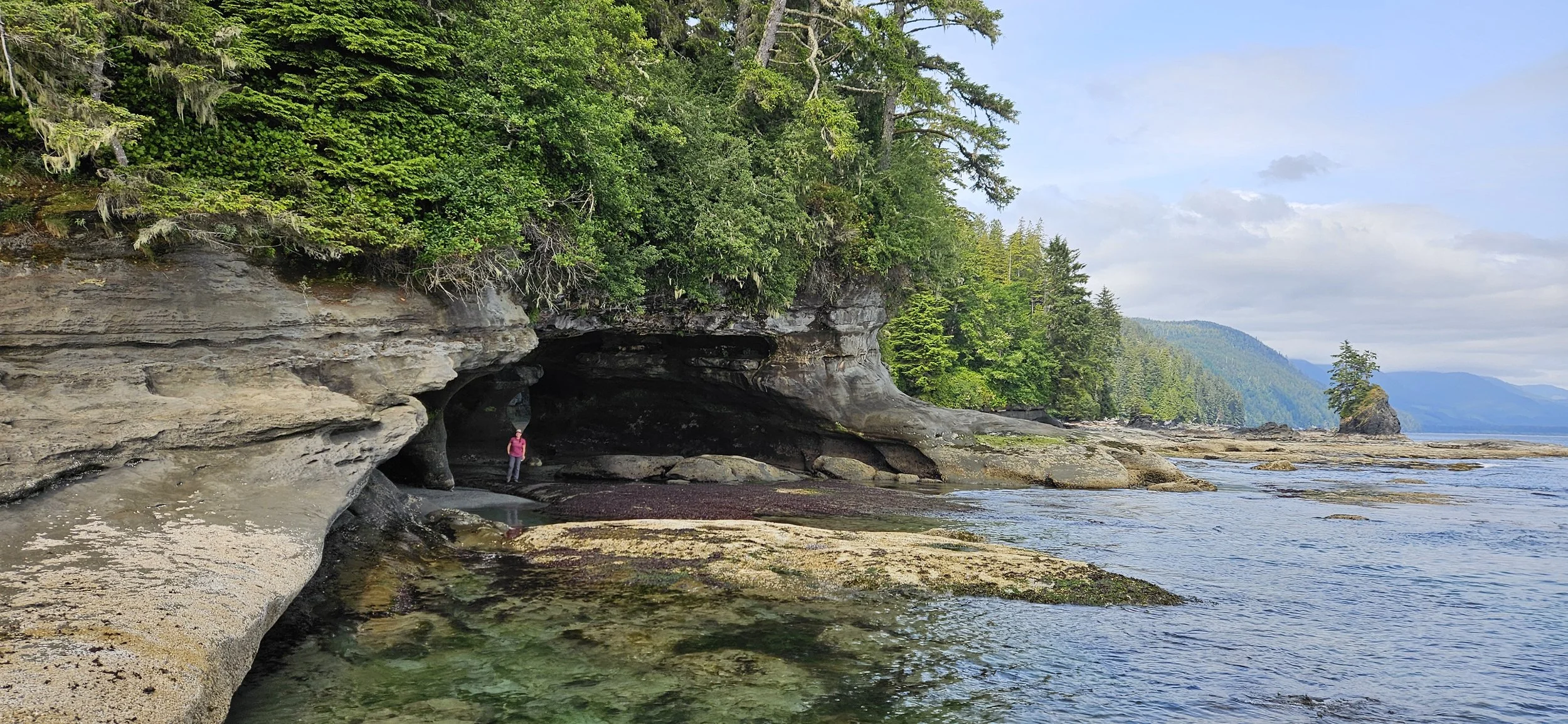 A person standing on a rocky shoreline near a rock formation with a cave, surrounded by lush green trees, with a view of the ocean and distant mountains under a partly cloudy sky.