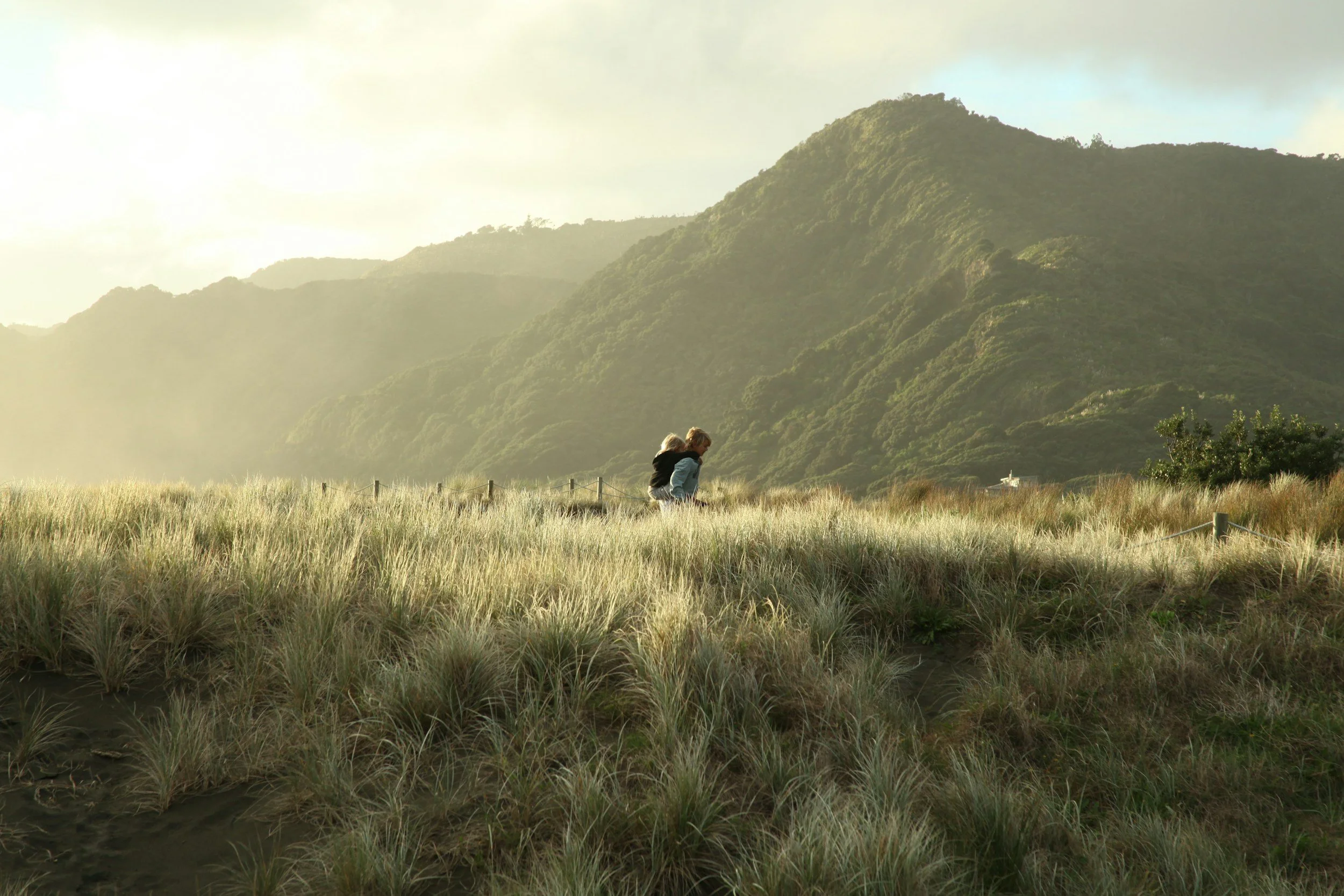 Two children sitting in tall grass with mountains in the background during a foggy or misty morning.