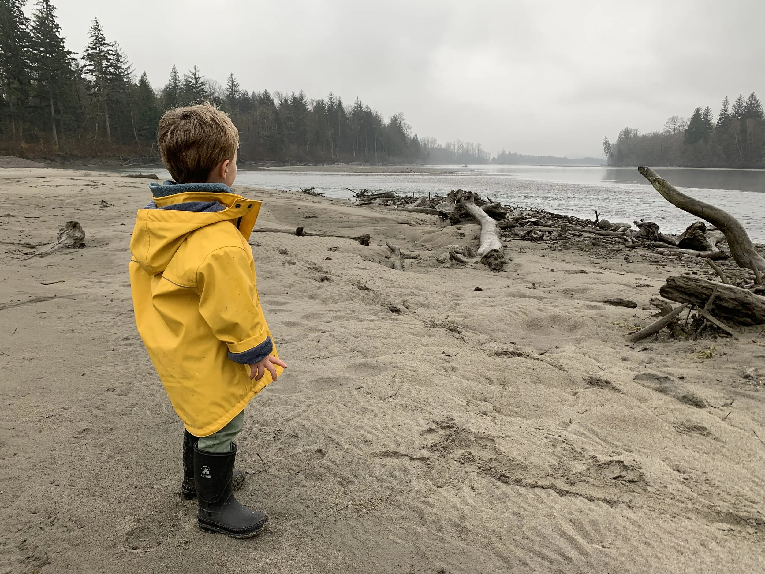 A young boy in a yellow raincoat and black rubber boots standing on a sandy beach by a river, looking out at the water with trees and overcast sky in the background.
