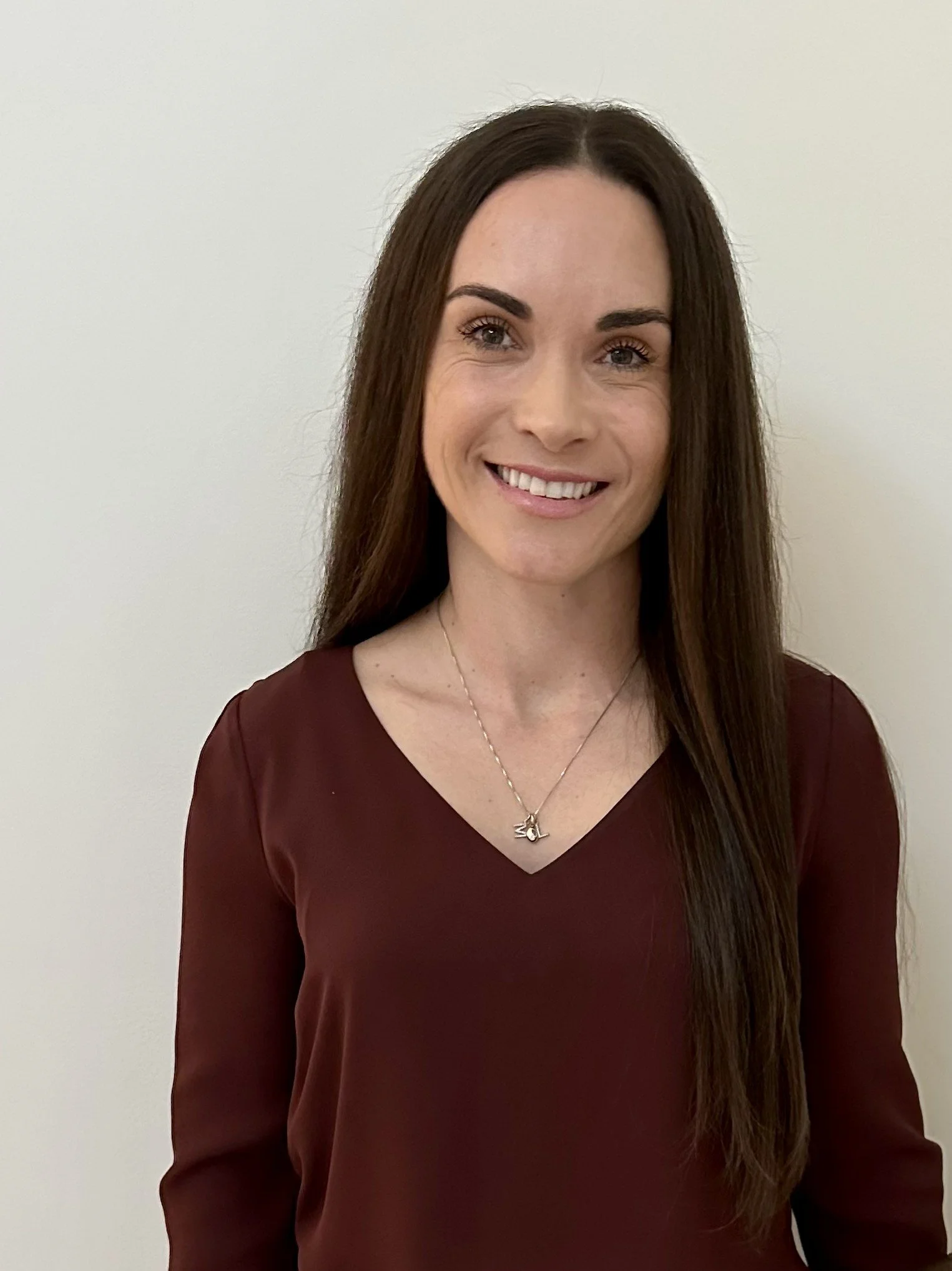 A woman with long brown hair, wearing a maroon long-sleeve top, standing against a plain light-colored background, smiling at the camera, wearing a necklace with a small pendant.
