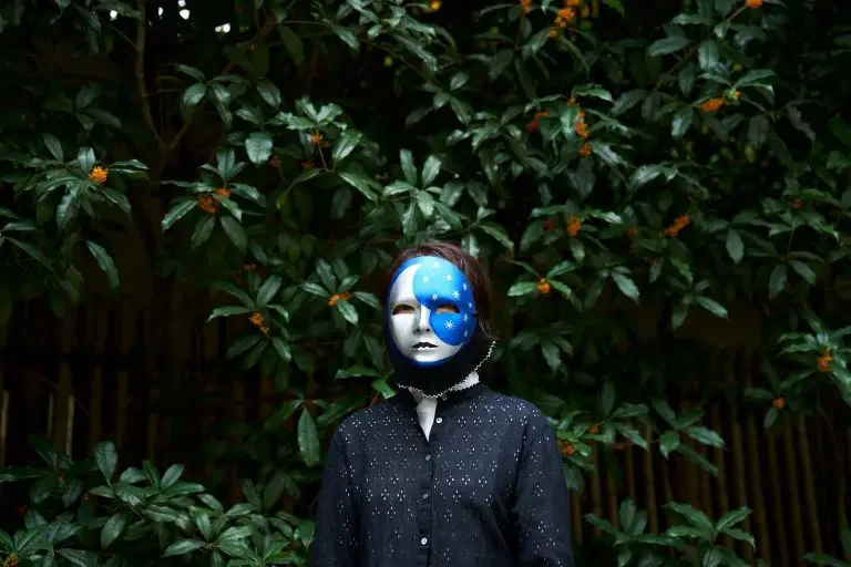 夜の森の中で、白い仮面をつけた人物が立っている。仮面は青と白の顔のデザインが施されている。背後は緑の葉っぱに囲まれている。