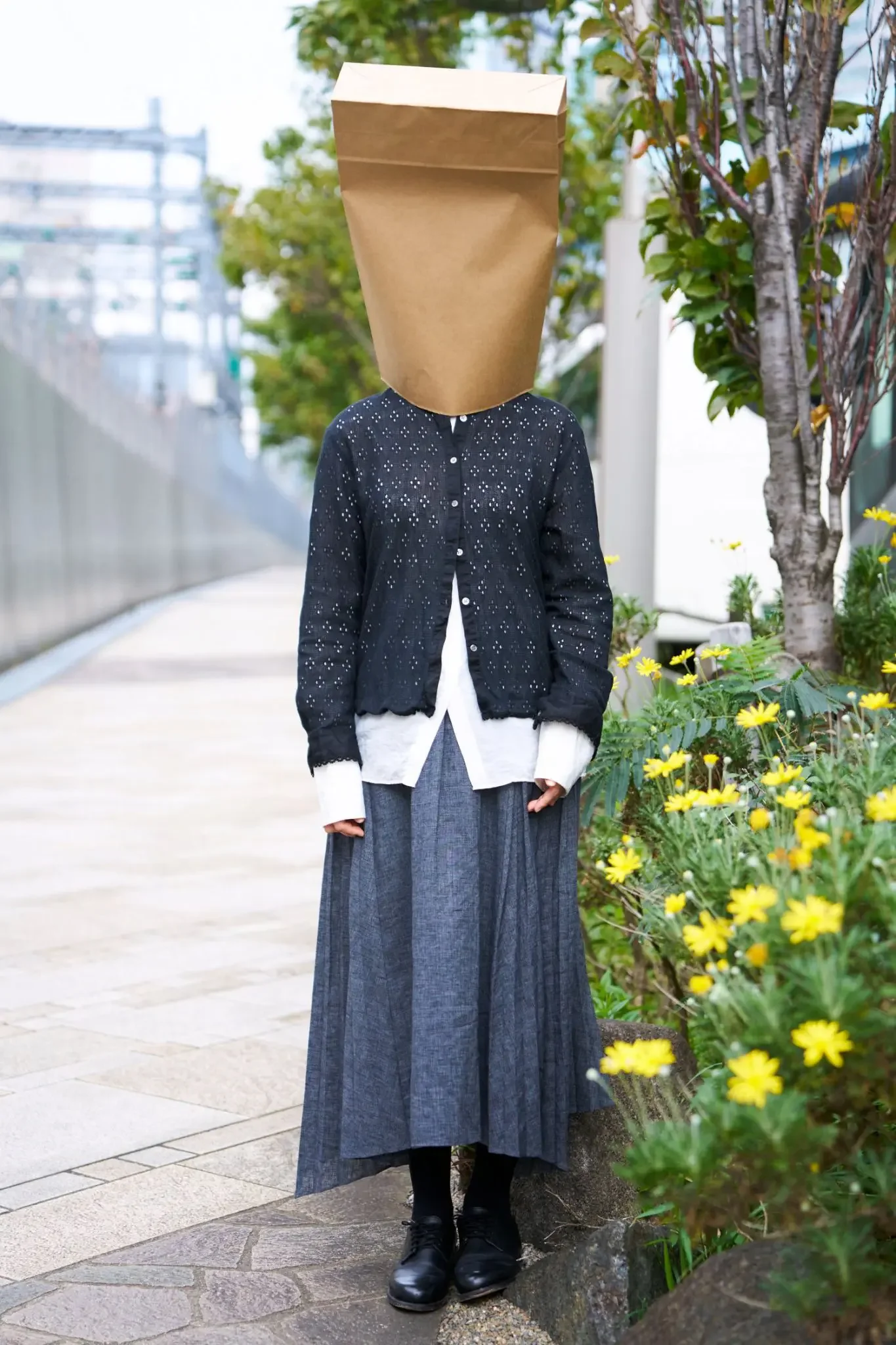 布袋をかぶった人物が街路に立っている。背景には木と花。